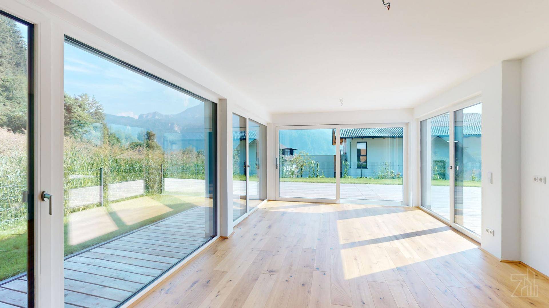 Heller, leerer Wohnraum mit großen Fensterfronten und Holzboden, der viel Tageslicht hereinlässt.