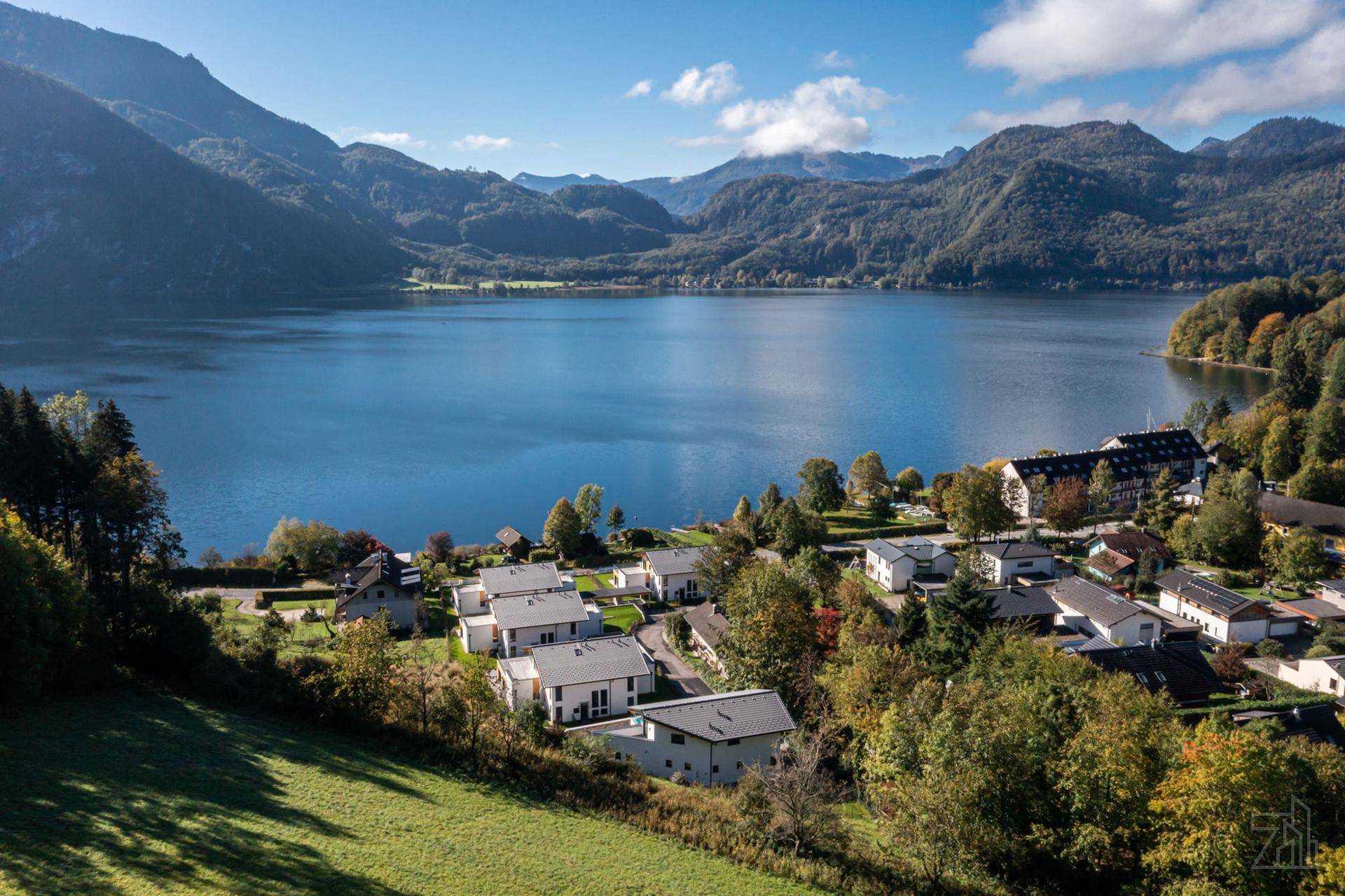 Malerische Luftaufnahme eines Dorfes am Seeufer, umgeben von Bergen und üppiger Natur.