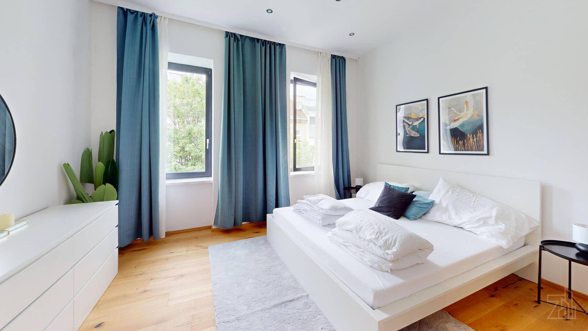 Geräumiges Schlafzimmer mit Holzboden, großen Fenstern und blauen Vorhängen.