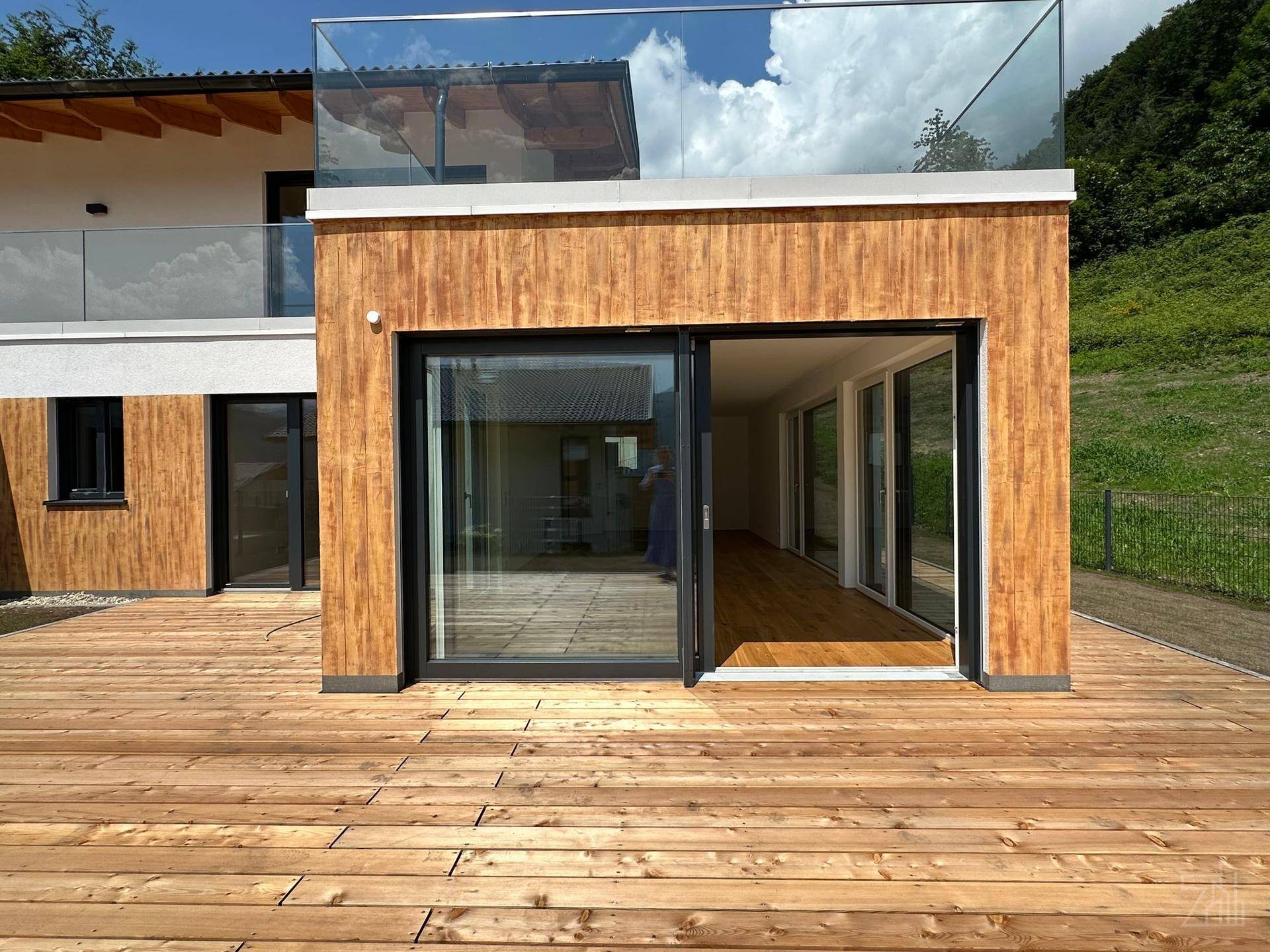 Geräumige Holzterrasse mit Glasgeländer und Zugang zum Innenbereich, ideal für sonnige Tage.