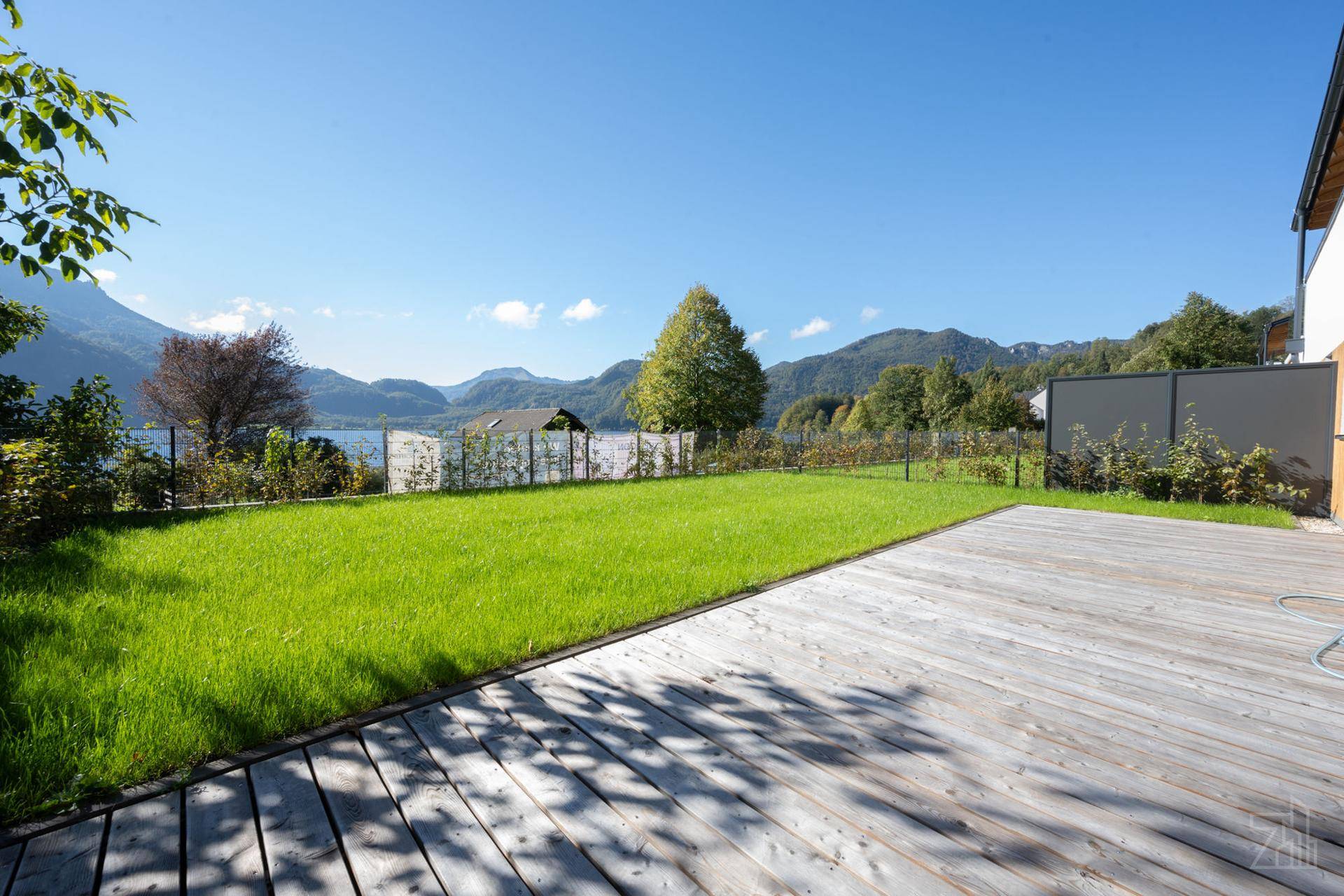 Weitläufige Rasenfläche und Holzterrasse mit Blick auf den See und die umliegende Natur.
