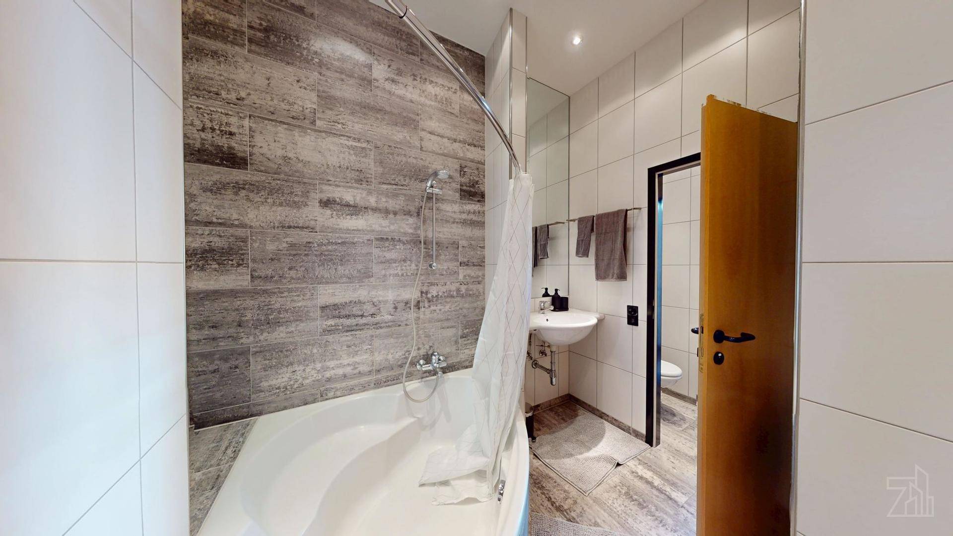 Modernes Badezimmer mit Badewanne, Dusche, weißen und grauen Fliesen sowie einem Waschbecken.
