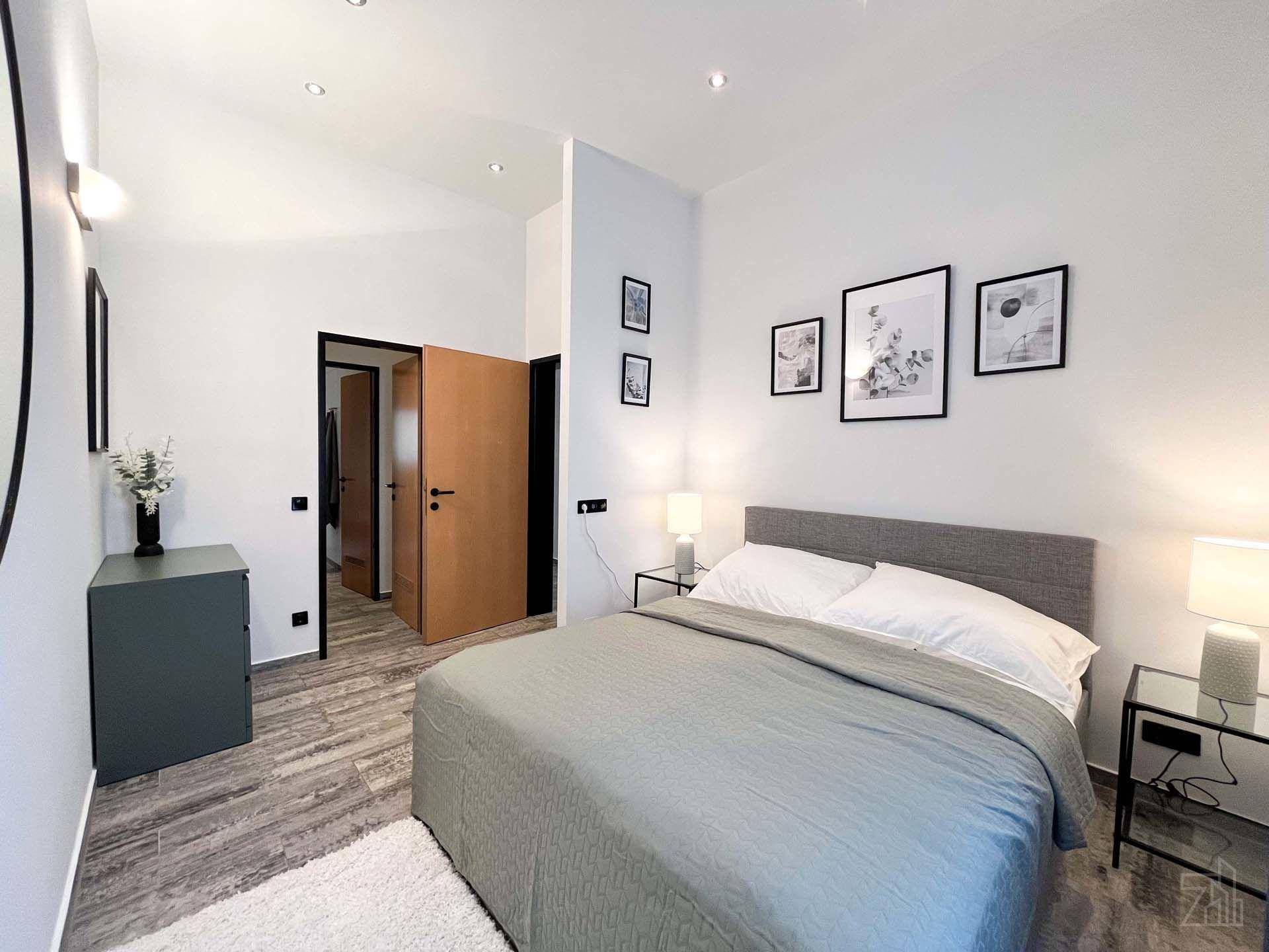 Helles Schlafzimmer mit grauem Bett, stilvollen Bildern an der Wand und Holzboden.