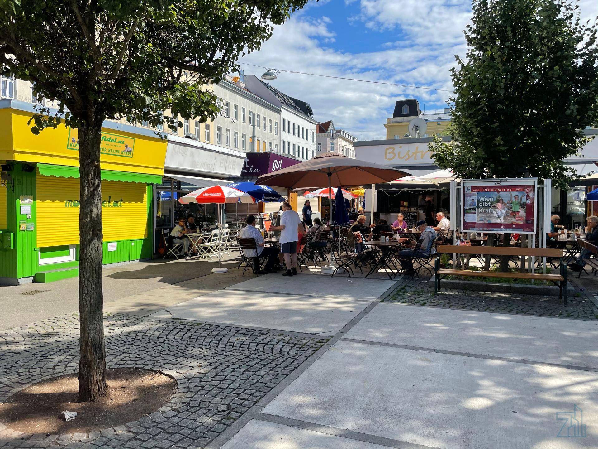 Lebhafter Außenbereich mit Cafés, Sonnenschirmen und Menschen an einem sonnigen Tag.