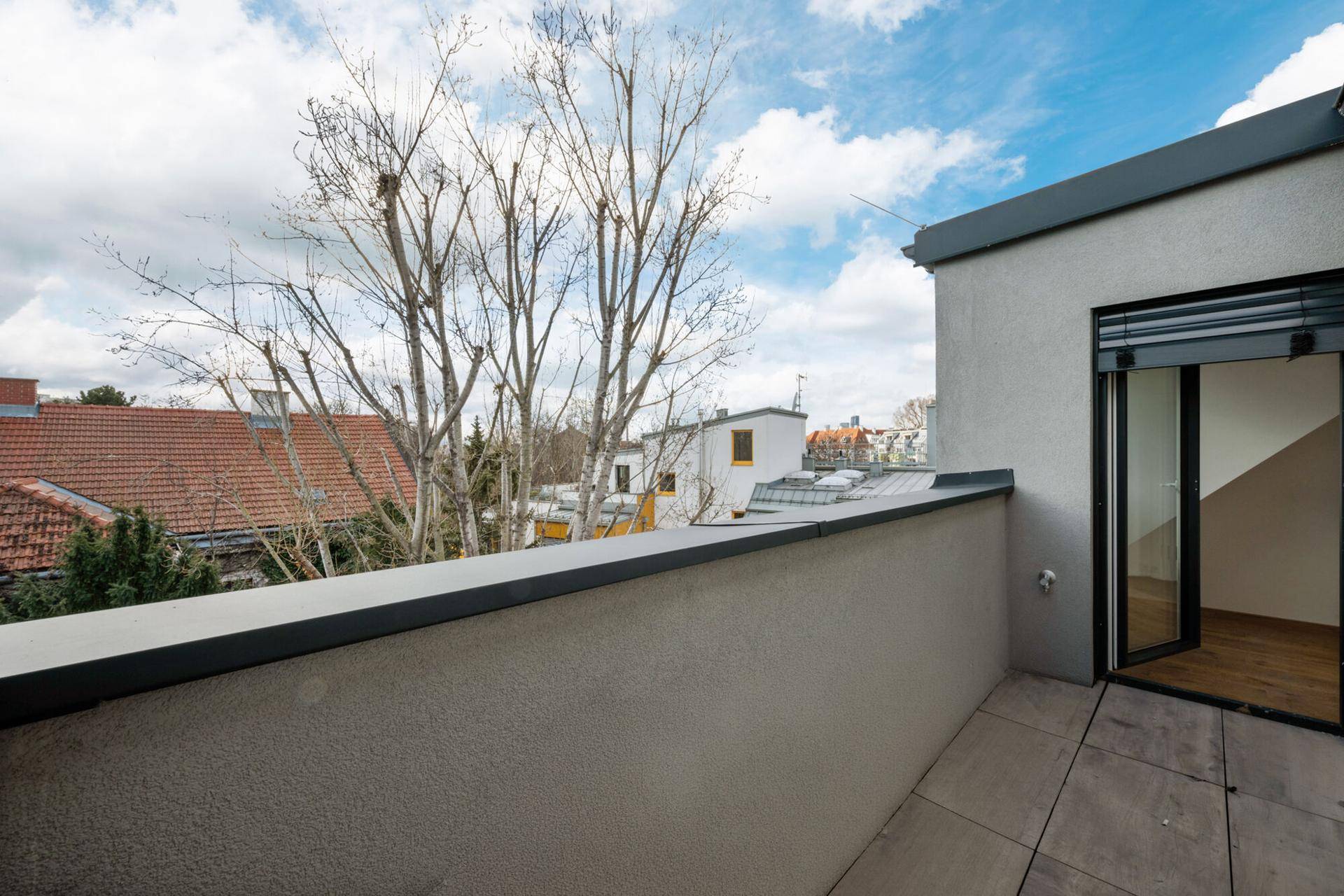 Terrasse mit Fliesenboden und Zugang zum Innenbereich, umgeben von Bäumen und anderen Gebäuden.