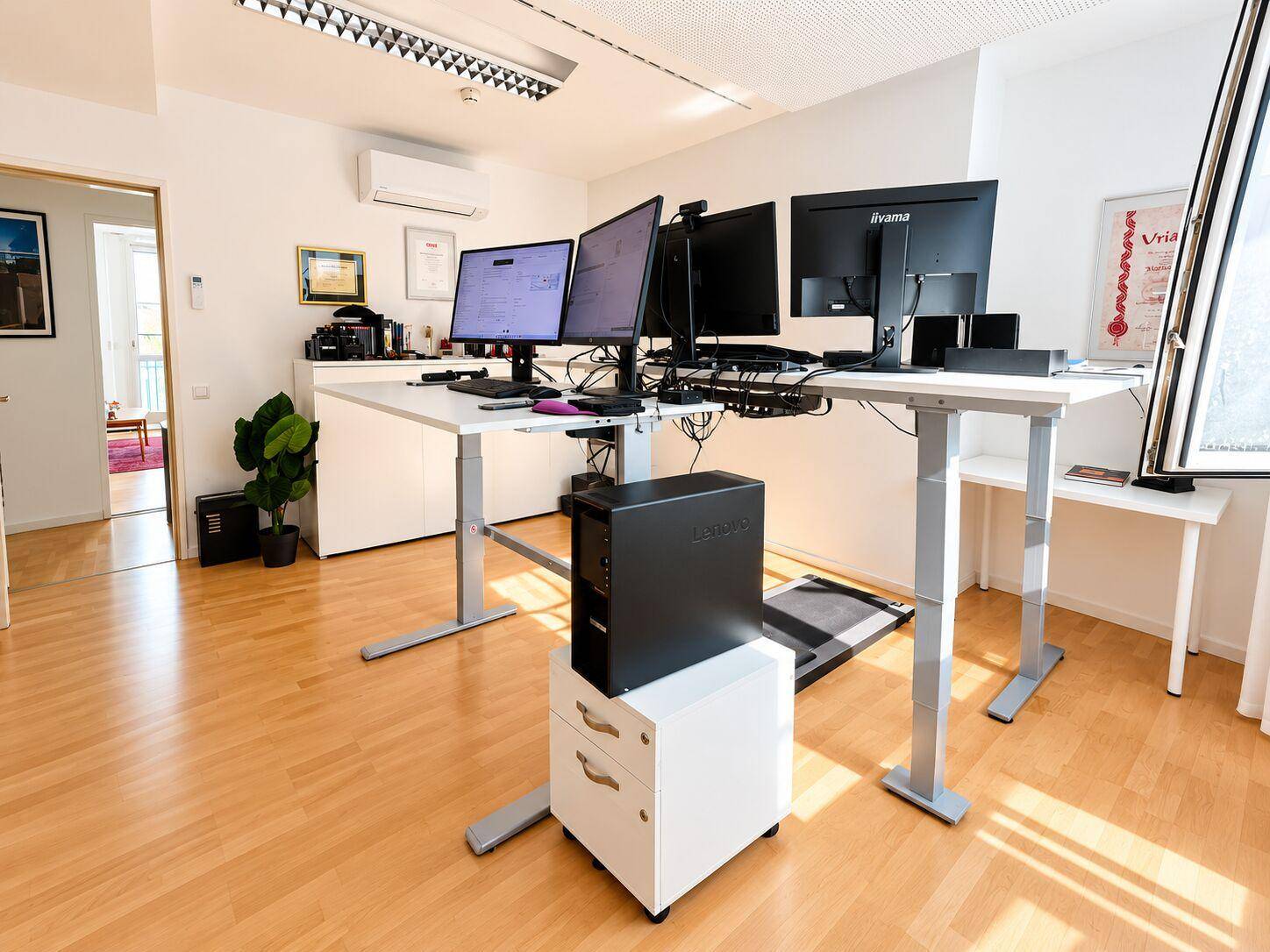 Geräumiges Büro mit mehreren Monitoren, Klimaanlage und Blick in einen angrenzenden Raum.