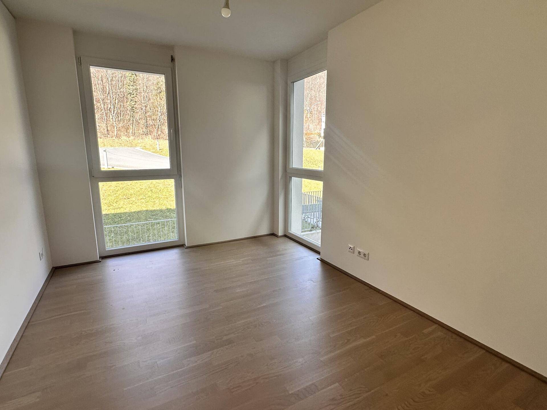 Helles Zimmer mit Parkettboden und zwei Fenstern mit Ausblick ins Grüne.