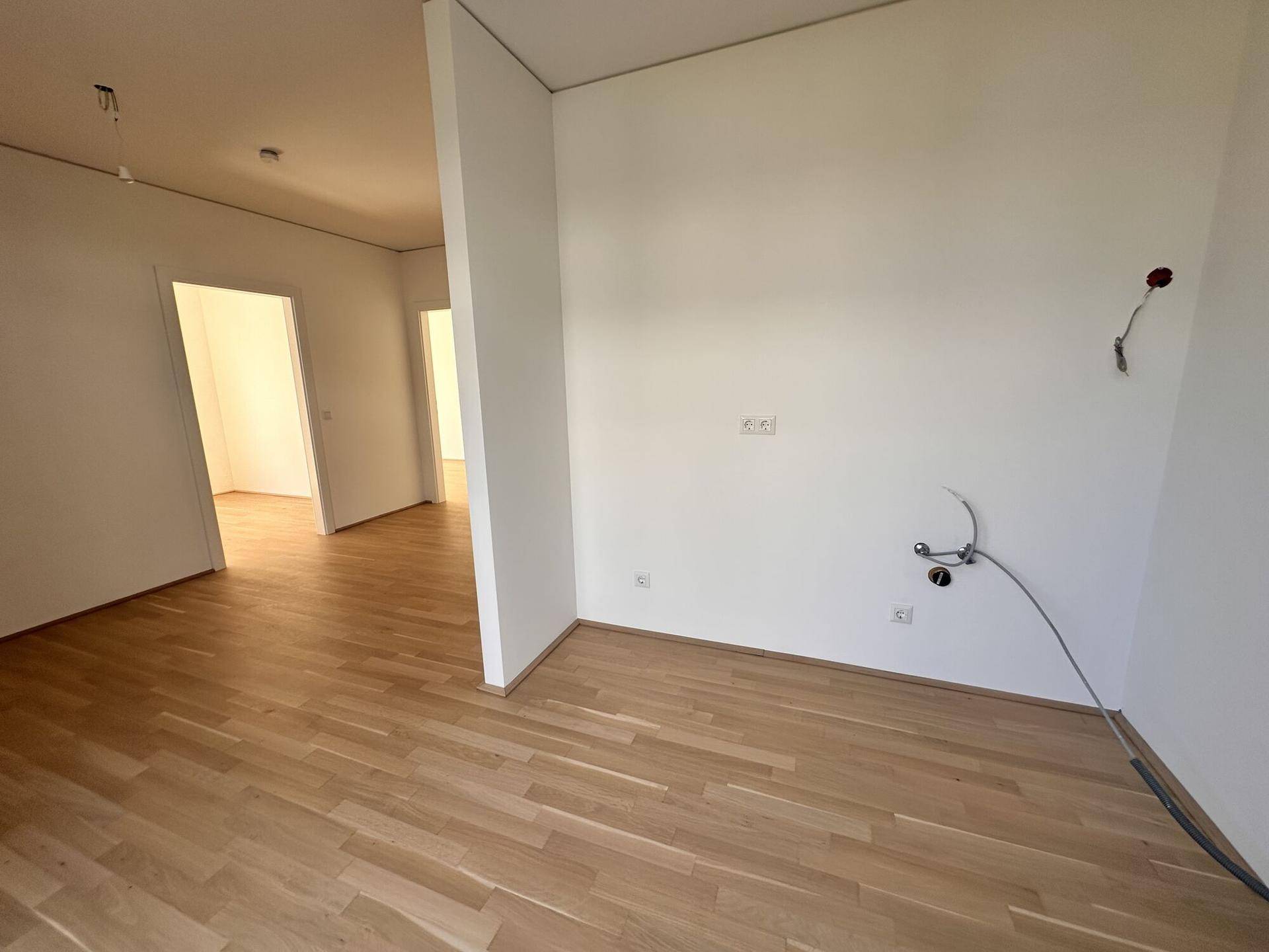 Leerer Raum mit hellem Holzparkettboden und vorbereiteten Anschlüssen für eine zukünftige Küche.