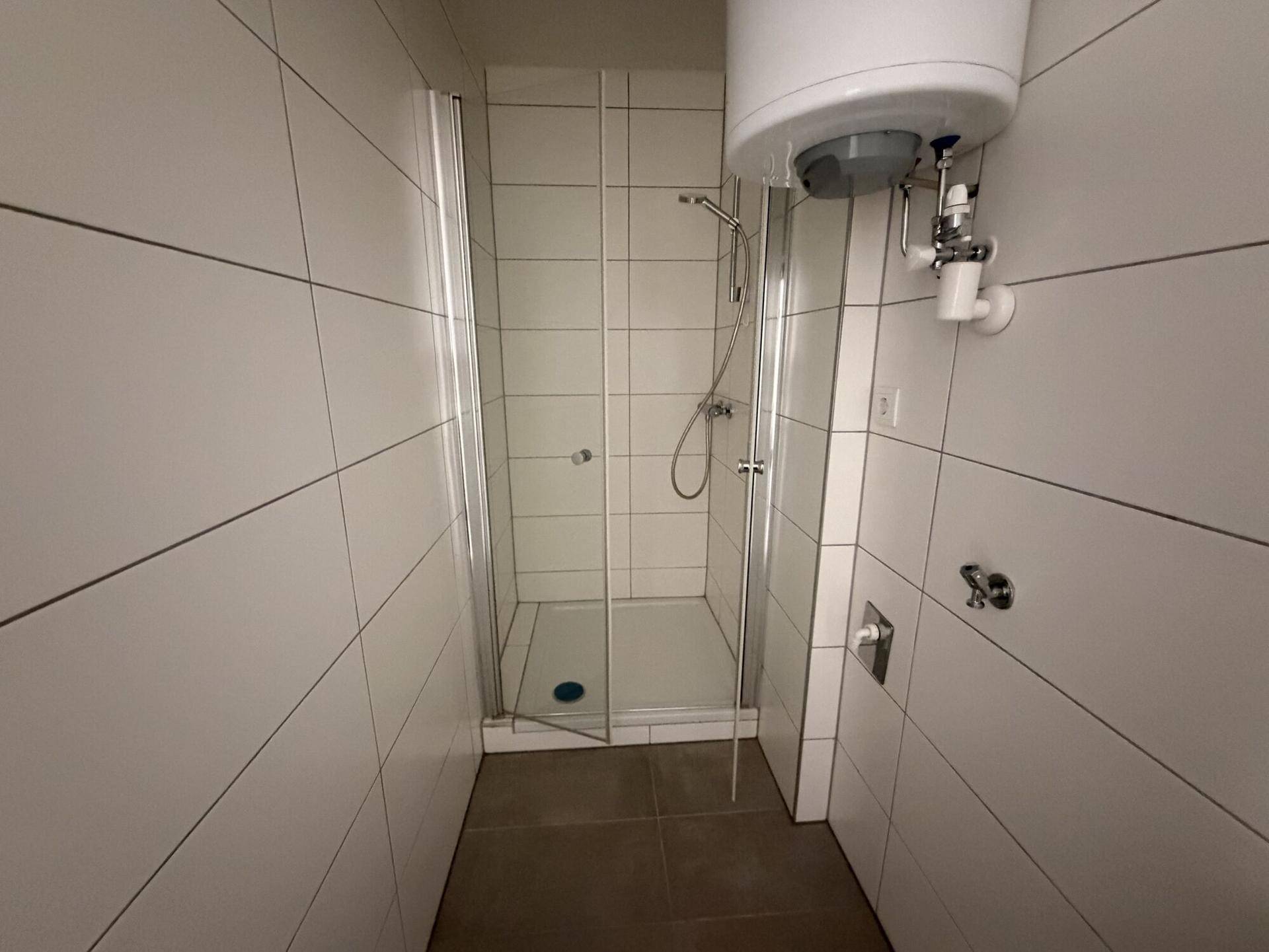 Modern geflieste Dusche mit Glasabtrennung und Warmwasserboiler in einem kleinen Bad.