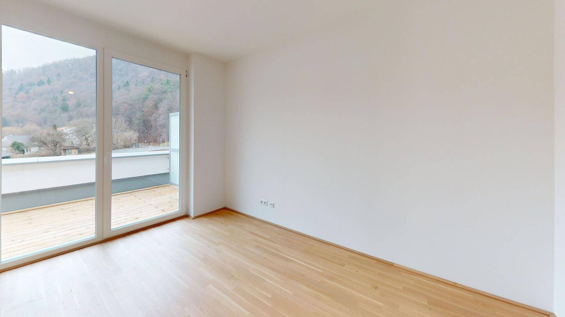 Lichtdurchfluteter Raum mit Holzboden und bodentiefen Fenstern, die einen Blick ins Grüne ermöglichen.