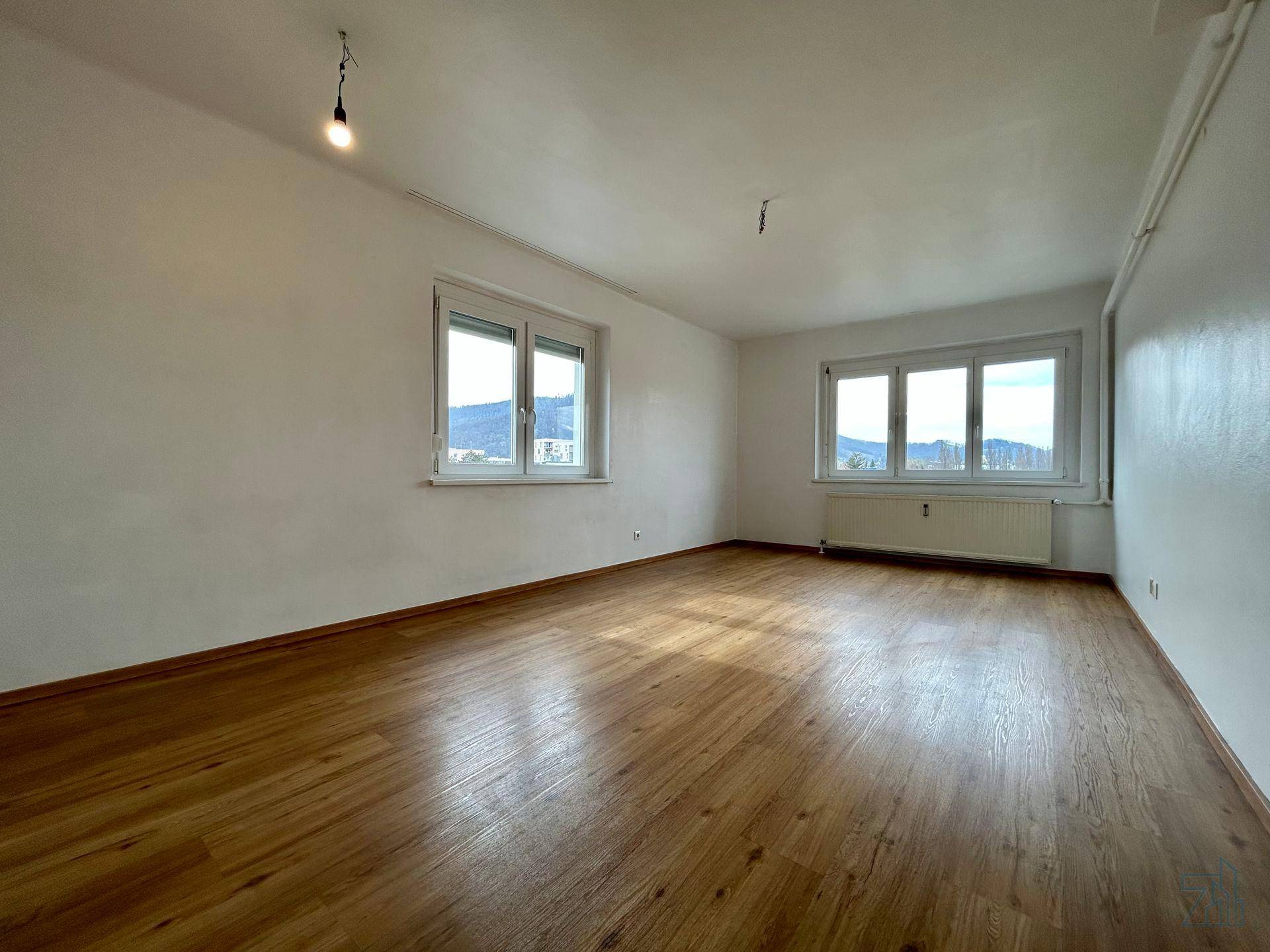 Heller, leerer Raum mit Holzfußboden und zwei Fenstern, die einen Blick auf die umliegenden Hügel bieten.