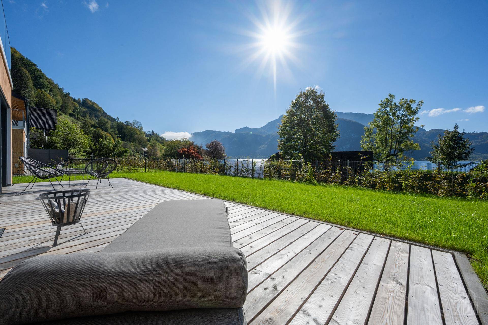 Sonnige Holzterrasse mit Liegestuhl und Gartenmöbeln, bietet einen weiten Blick auf den See und die Berge.