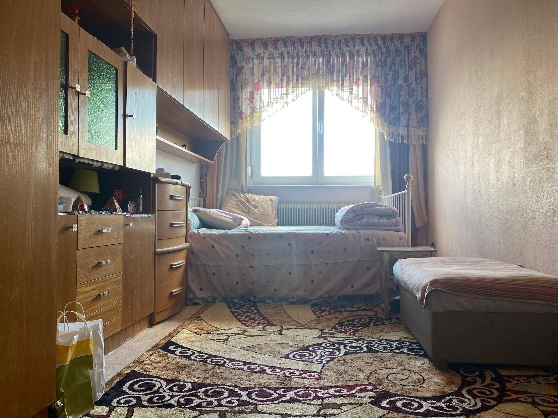 Gemütliches Schlafzimmer mit Holzmöbeln, einem Einzelbett und einem Fenster mit floralen Vorhängen.