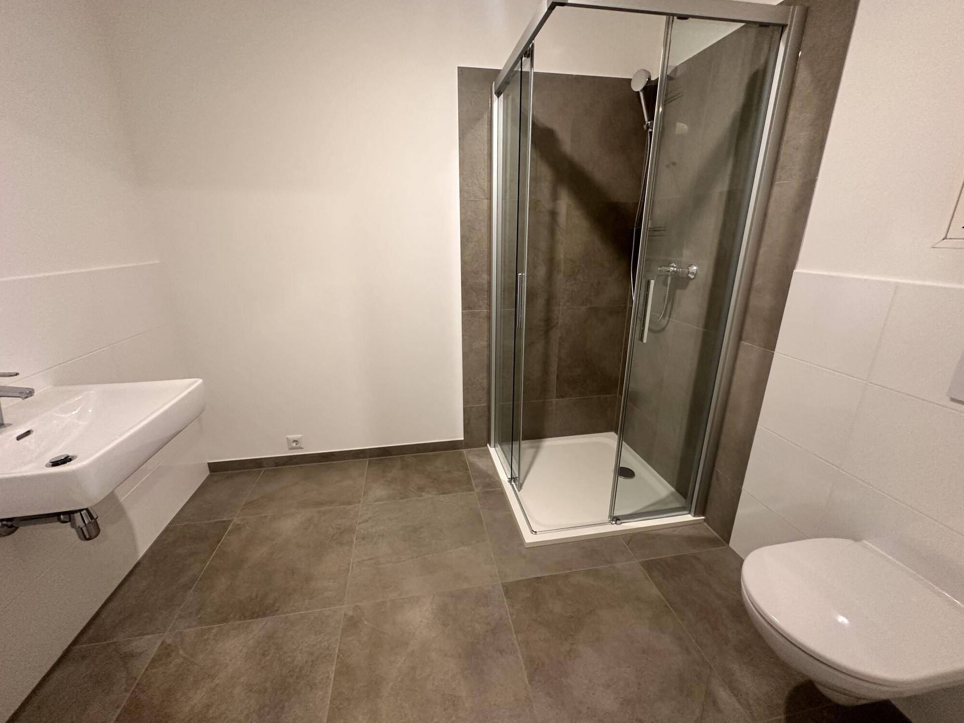 Modernes Badezimmer mit grauen Fliesen, einer Glasduschkabine, einem Waschbecken und einem wandhängenden WC.