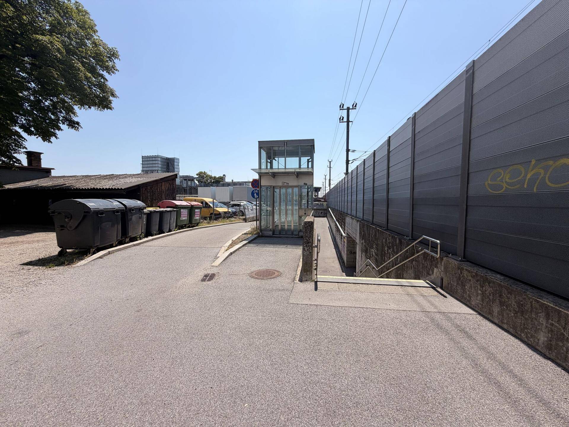 Außenbereich mit Weg, einem Aufzug und einer Lärmschutzwand entlang der Bahngleise.