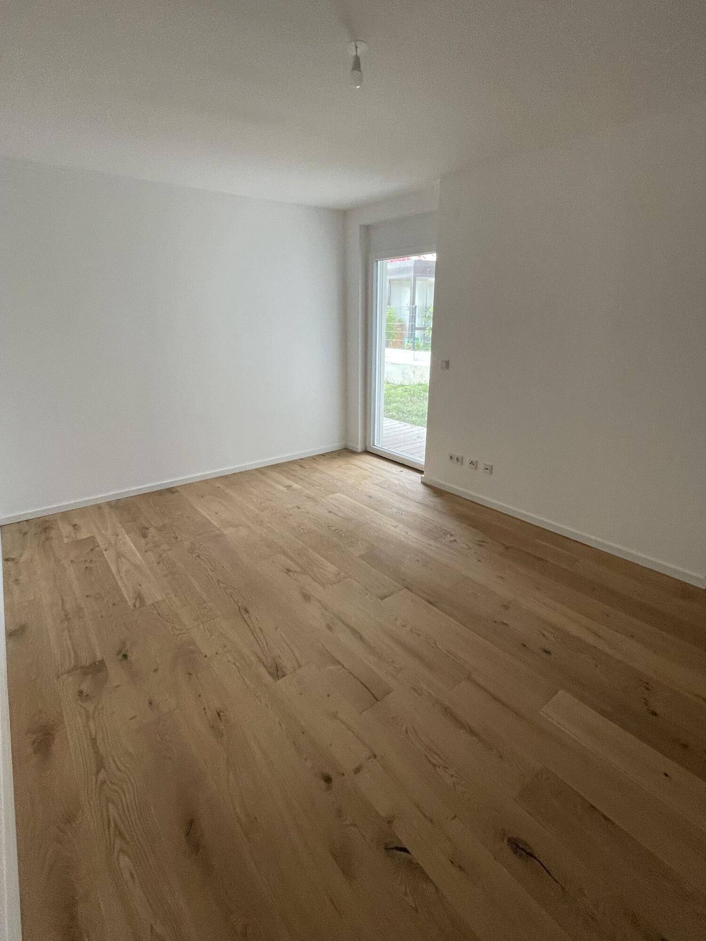 Leerer Raum mit hellem Holzparkettboden, weißen Wänden und Zugang zu einem Außenbereich.