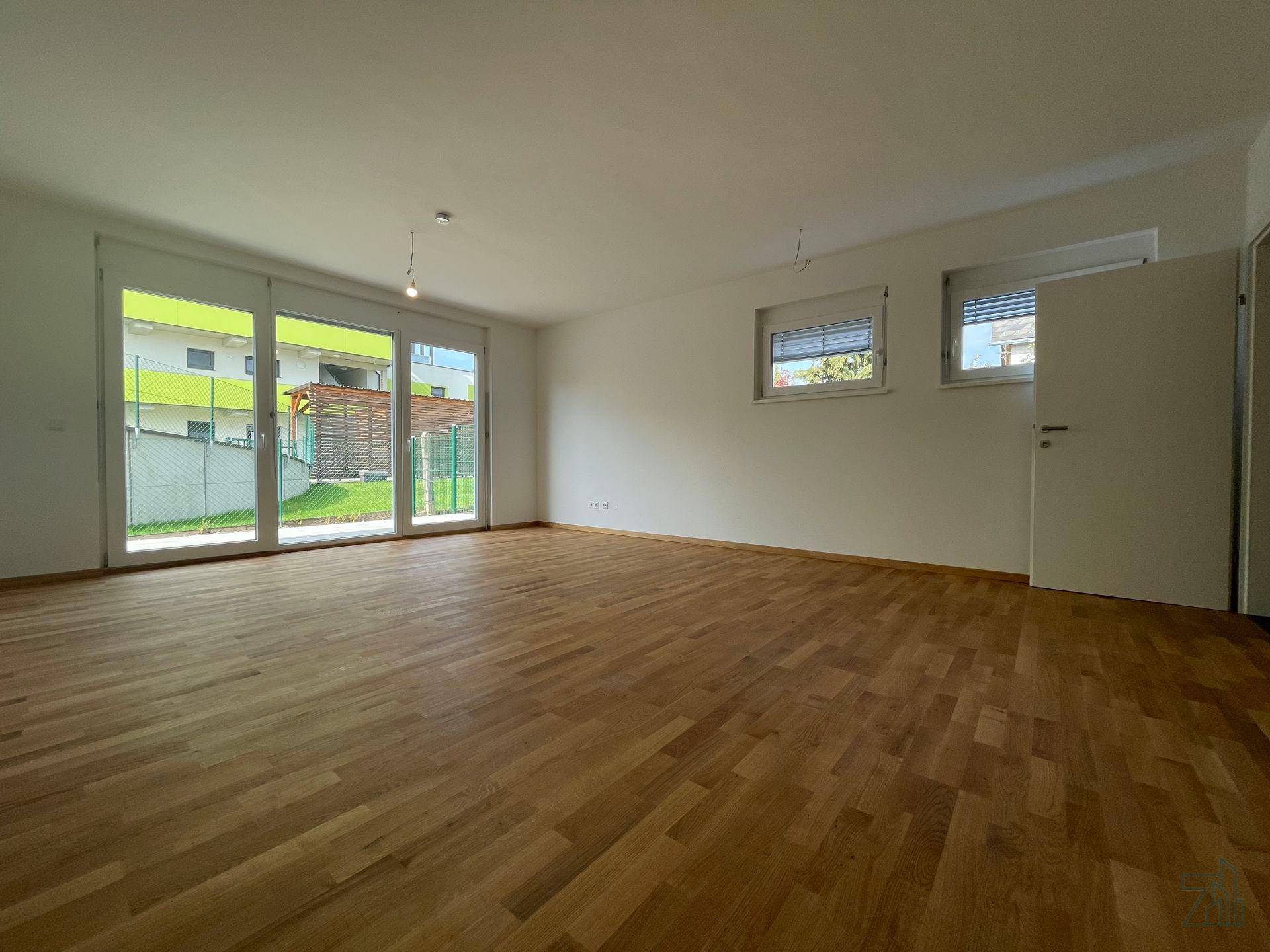 Helles, leeres Wohnzimmer mit Holzboden und großen Glastüren, die zum Garten führen.