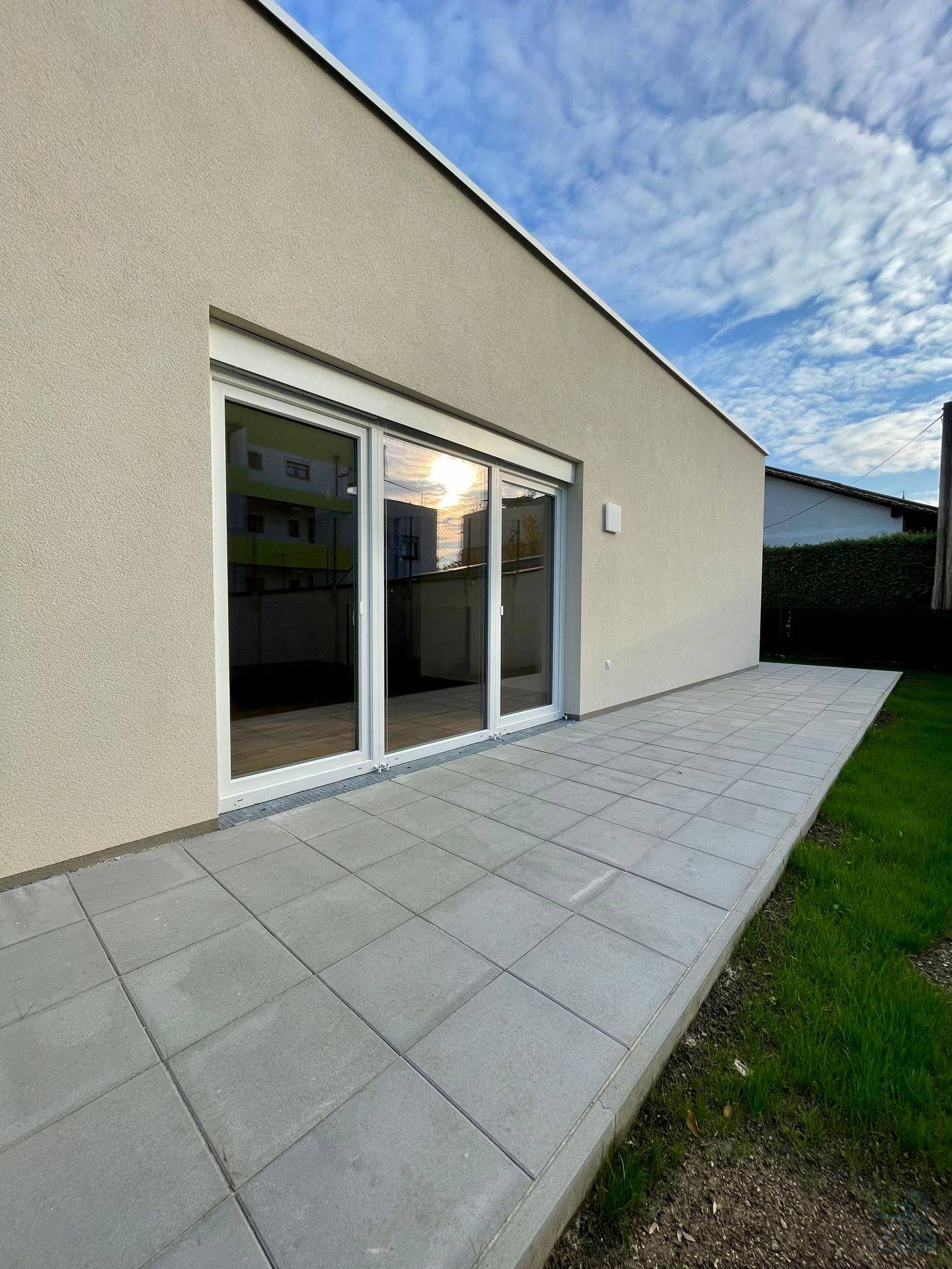 Seitenansicht des Gebäudes mit großer Glasschiebetür und der angrenzenden gepflasterten Terrasse.