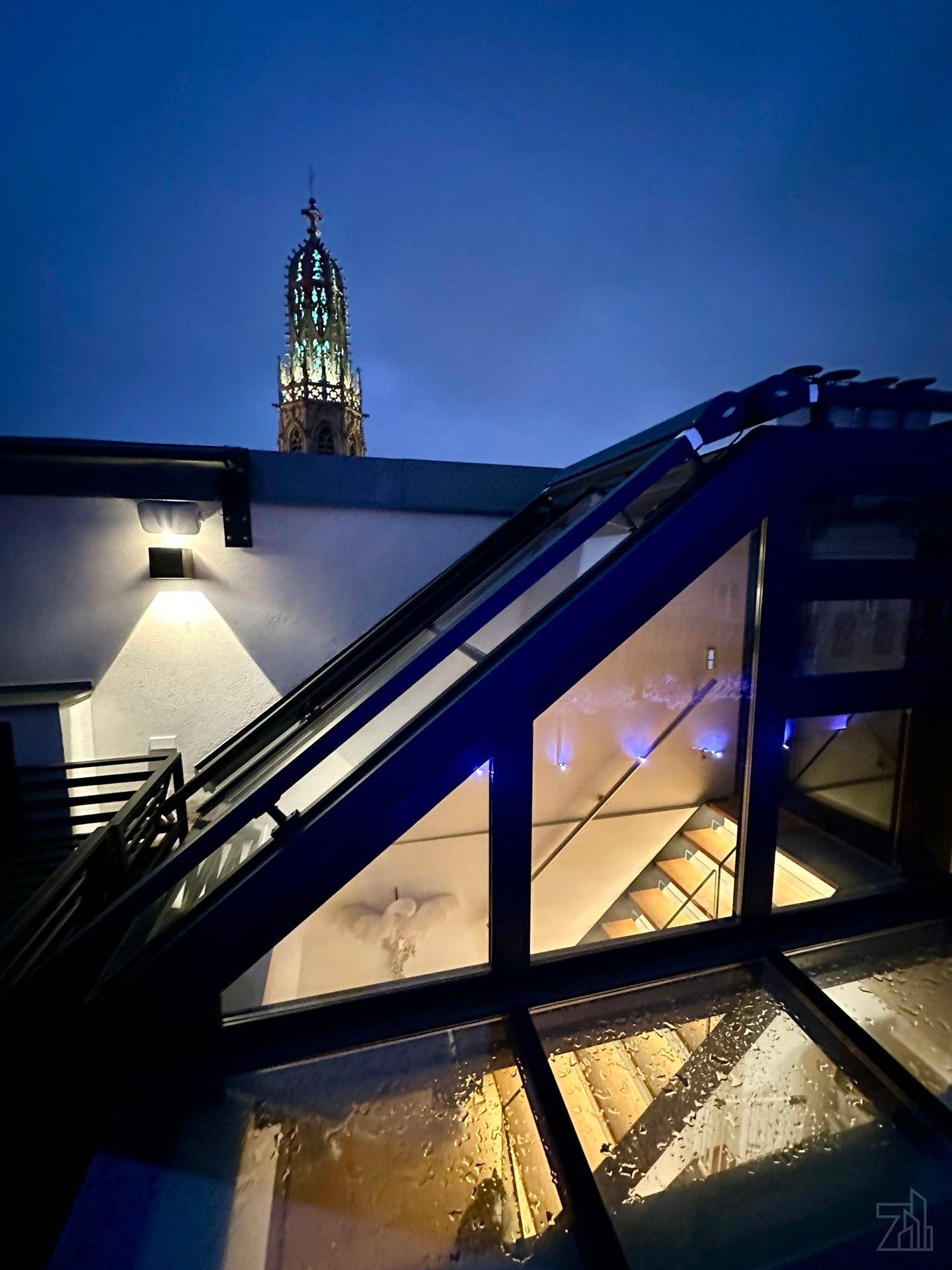 Glasstruktur auf dem Dach bei Nacht mit beleuchtetem Kirchturm im Hintergrund.