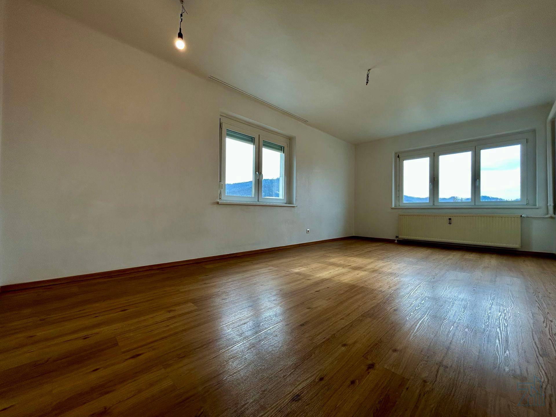 Heller, leerer Raum mit Holzboden und zwei Fenstern, die viel Tageslicht hereinlassen.