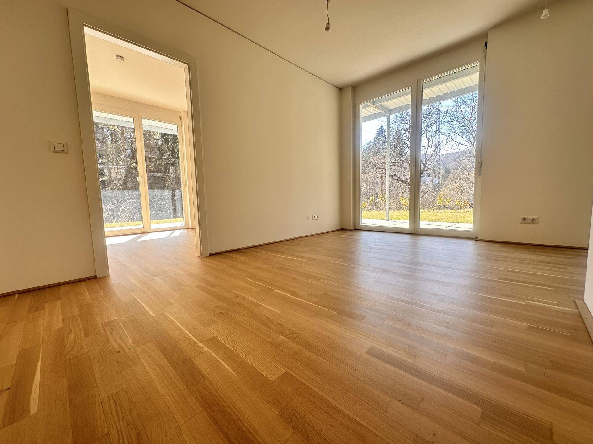Heller, leerer Raum mit Holzboden und großen Fenstern mit Blick ins Grüne.