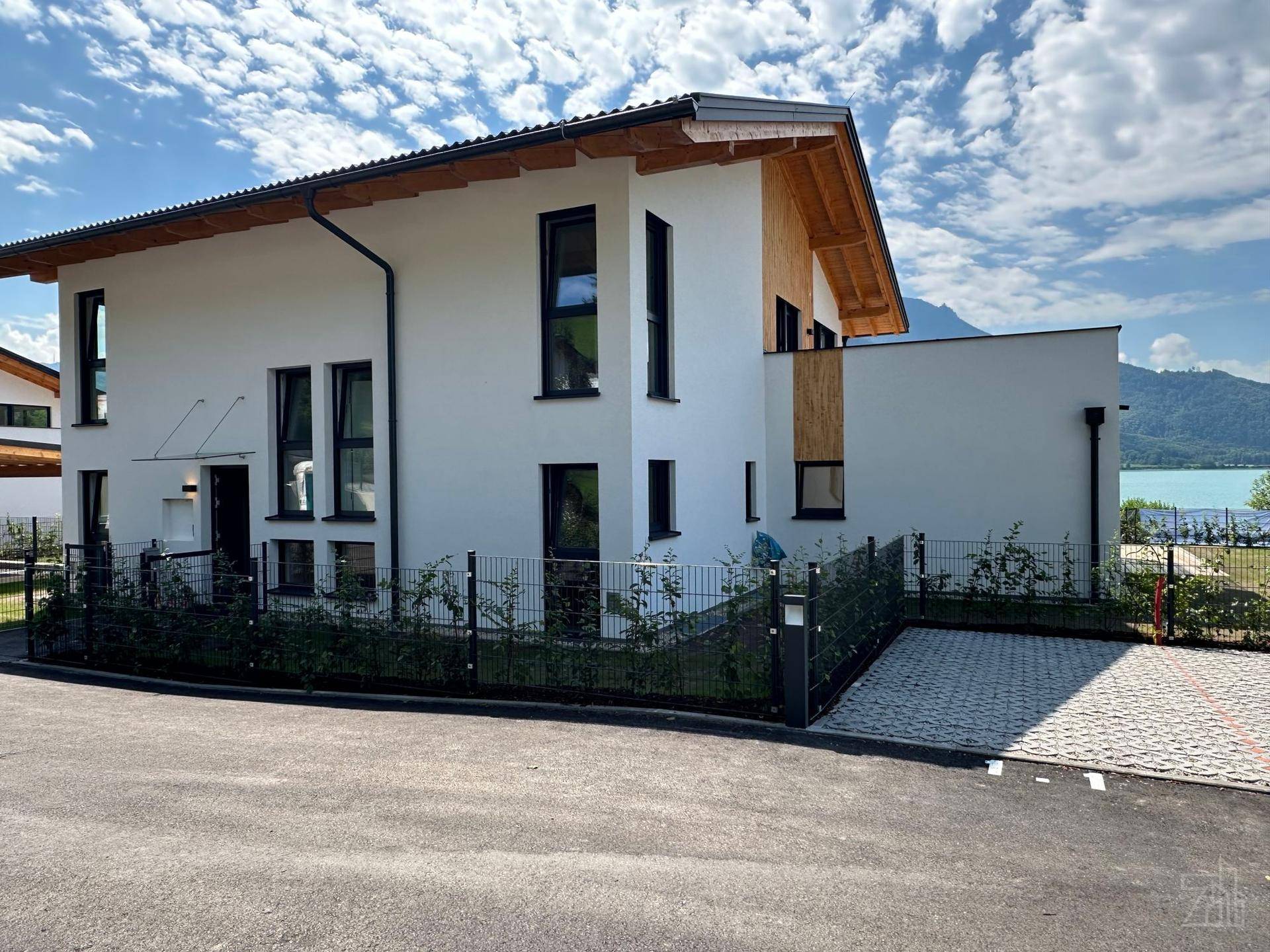 Modernes weißes Haus mit dunklen Fensterrahmen und Holzdachüberstand, mit Seeblick im Hintergrund.