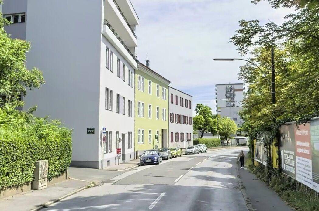 Straßenansicht mit Gebäuden, parkenden Autos und viel Grün entlang der Fahrbahn.
