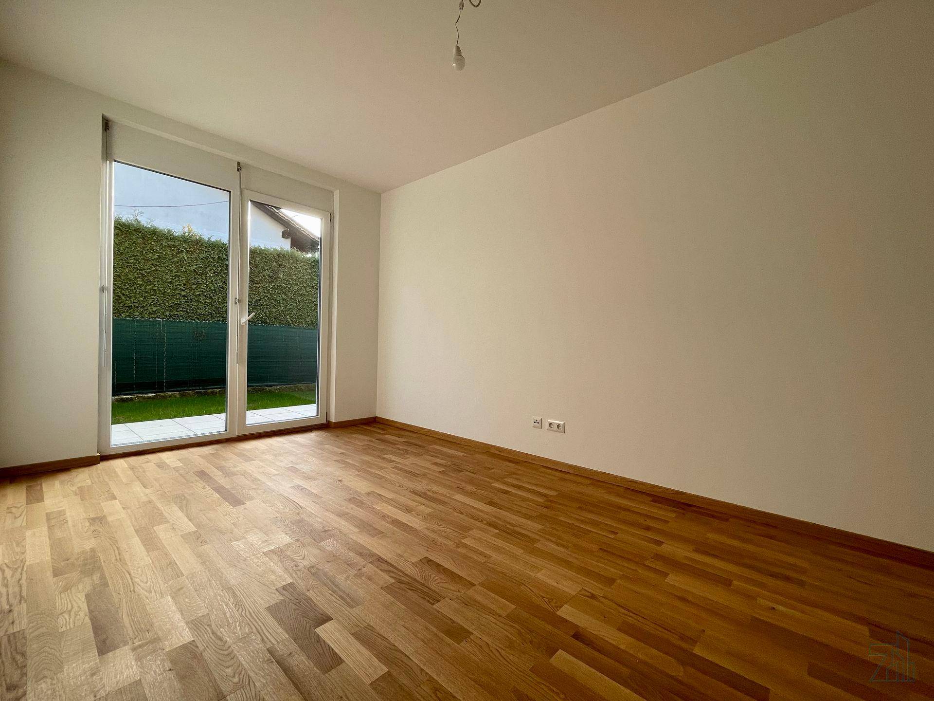 Leeres Zimmer mit Holzboden und großer Glastür, die zu einem von Hecken umgebenen Garten führt.