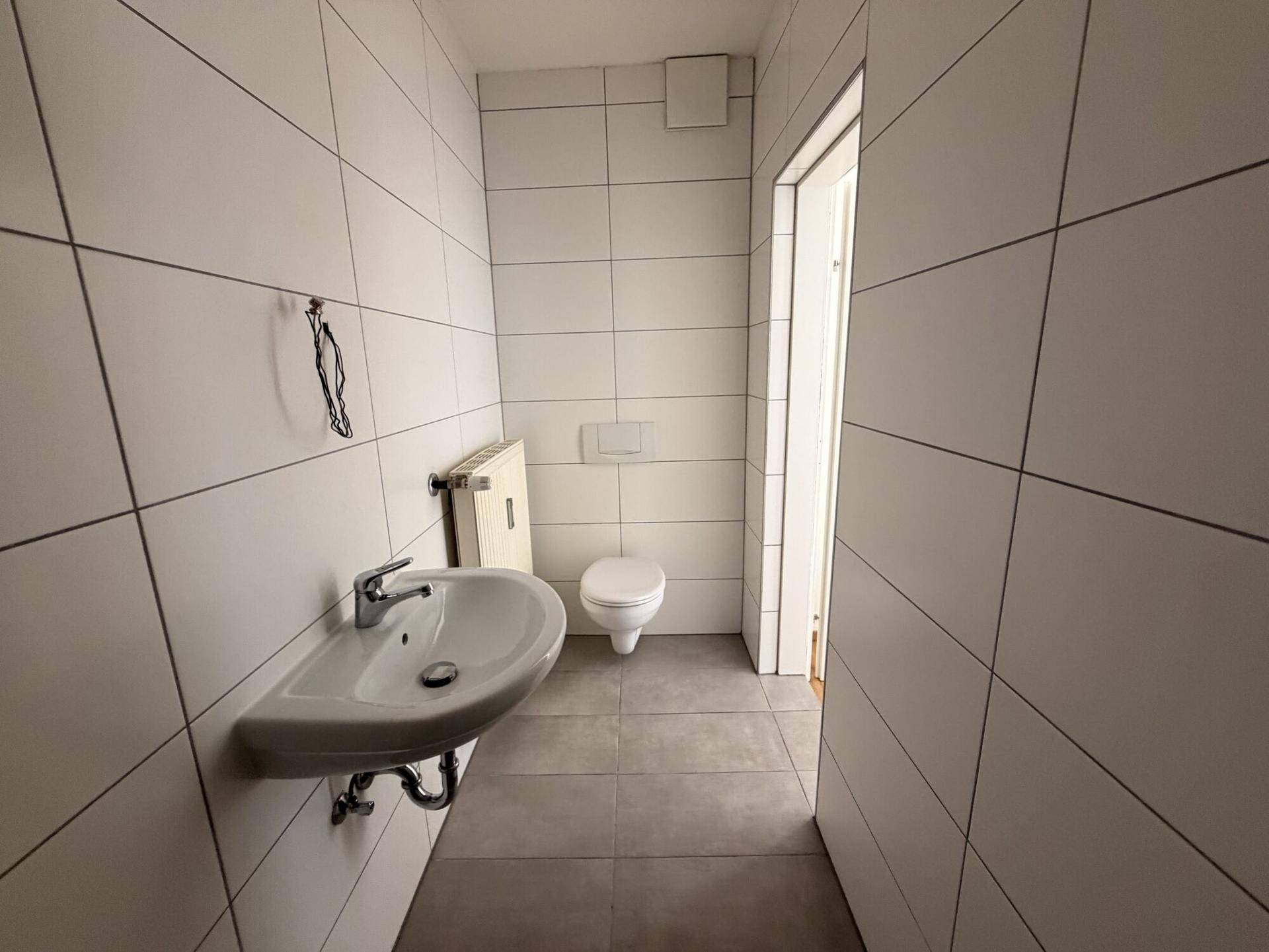 Helles WC mit Wand-WC, Waschbecken und Heizkörper, komplett gefliest.