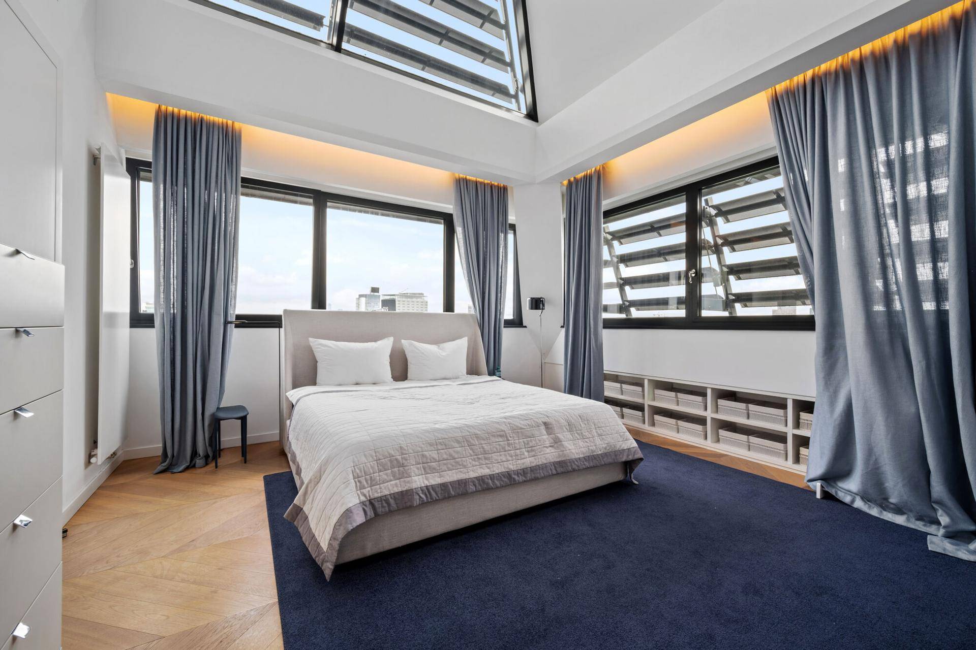 Geräumiges Schlafzimmer mit großem Bett, blauem Teppich und vielen Fenstern mit Stadtblick.