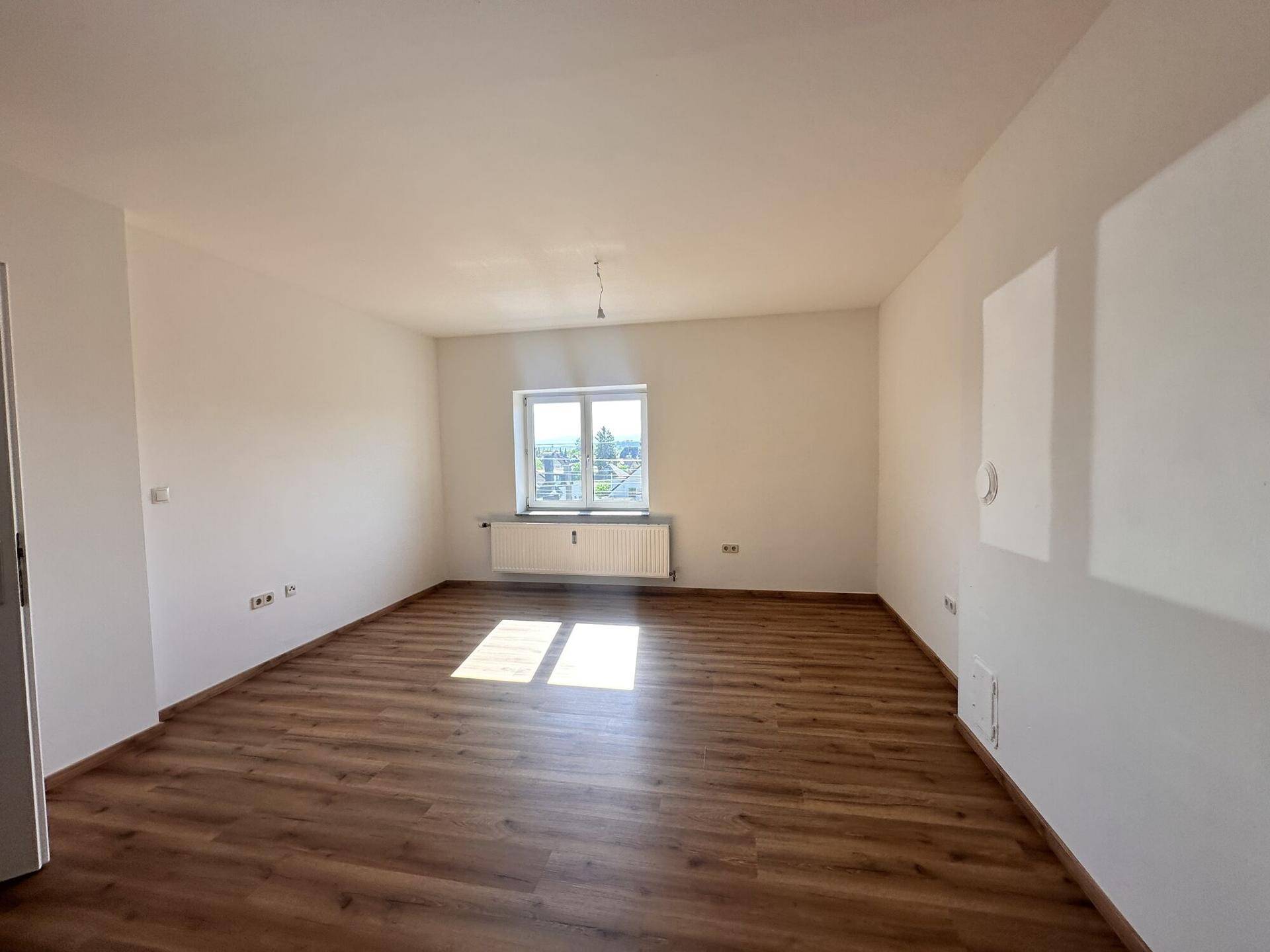 Leeres Wohnzimmer mit Laminatboden, weiß gestrichenen Wänden und einem großen Fenster.