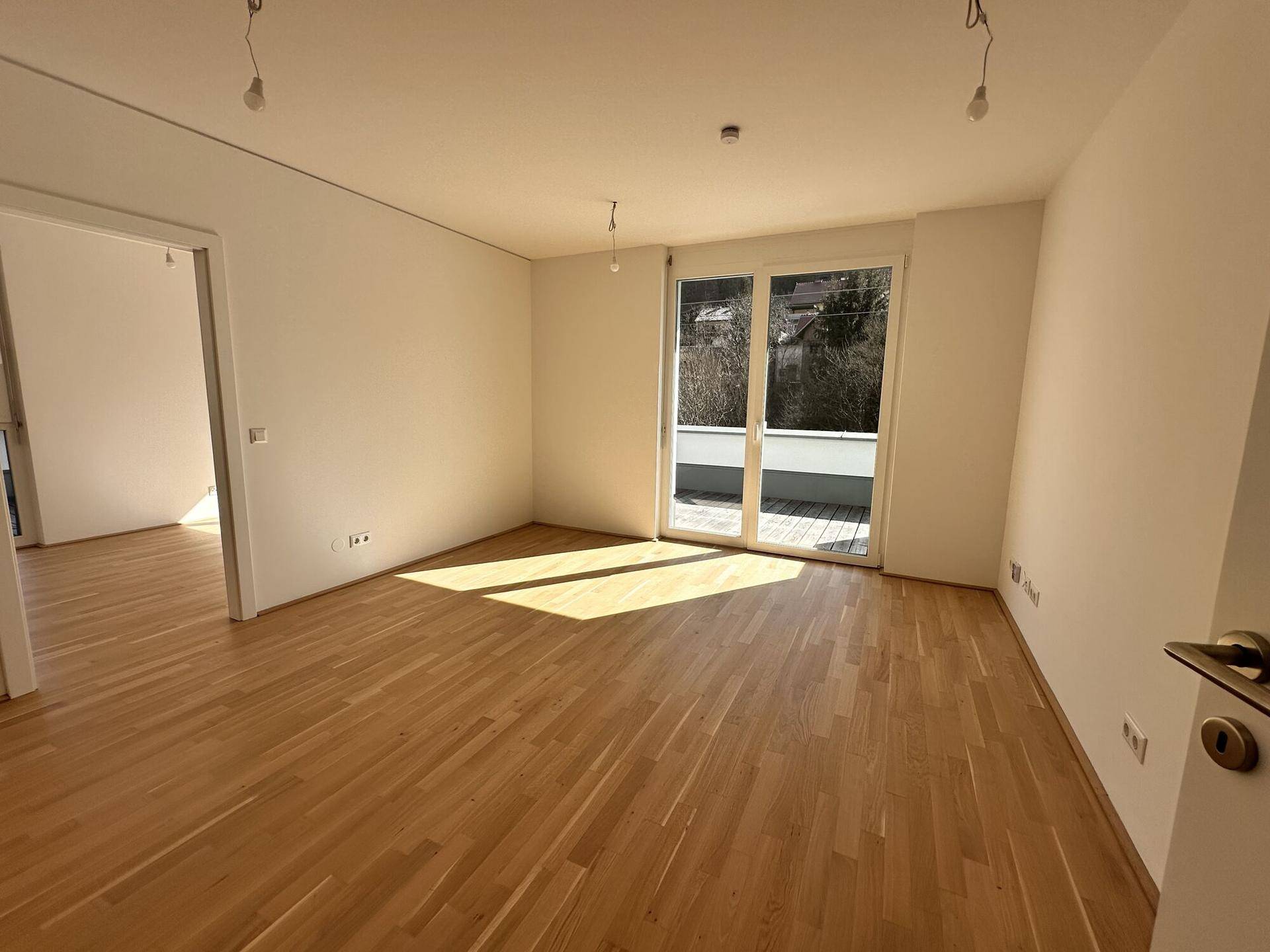 Heller Wohnbereich mit Parkettboden und Zugang zu einem weiteren Raum sowie zur Terrasse.
