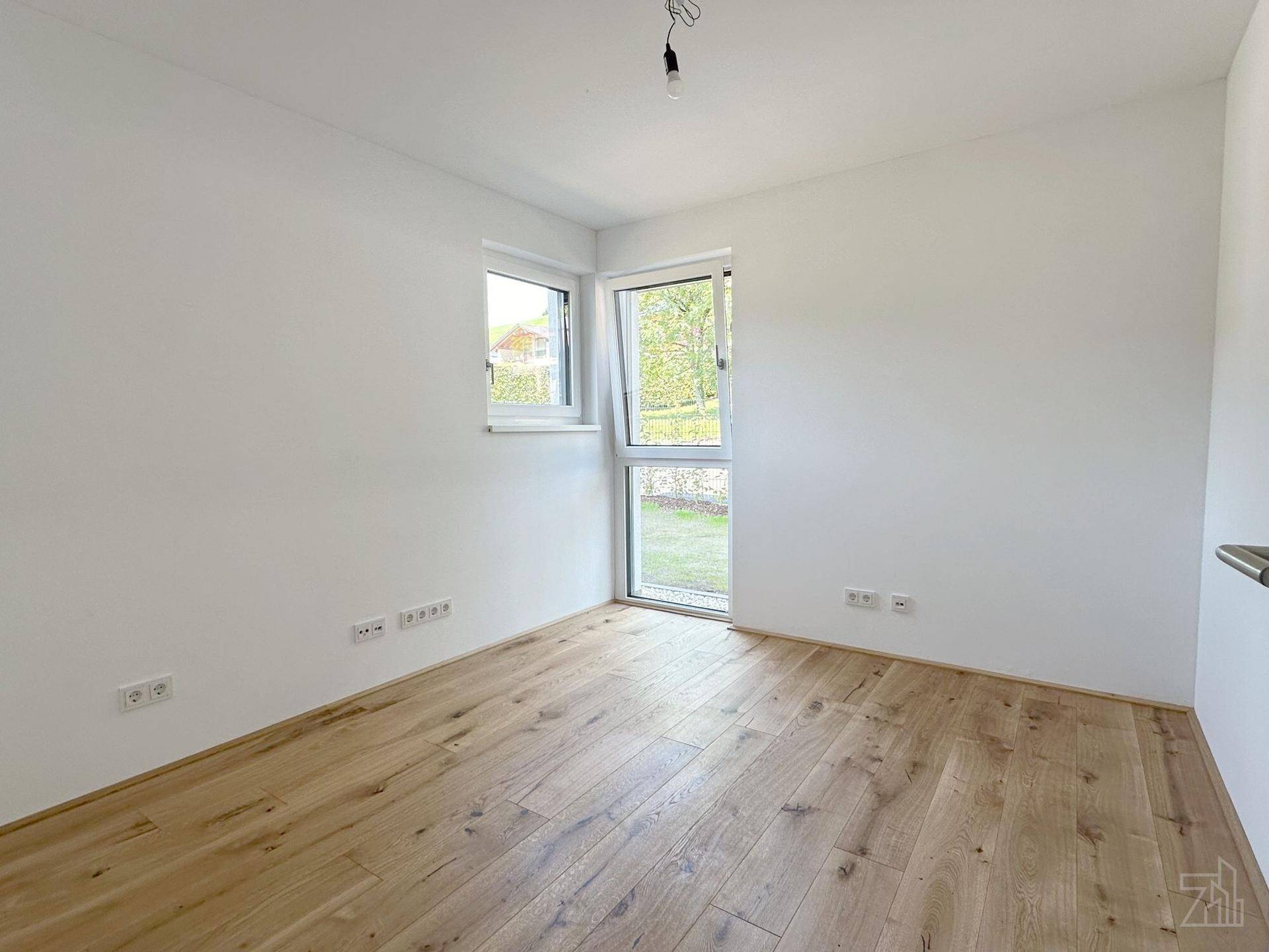 Helles Zimmer mit Holzboden und zwei Fenstern, die viel Tageslicht hereinlassen.