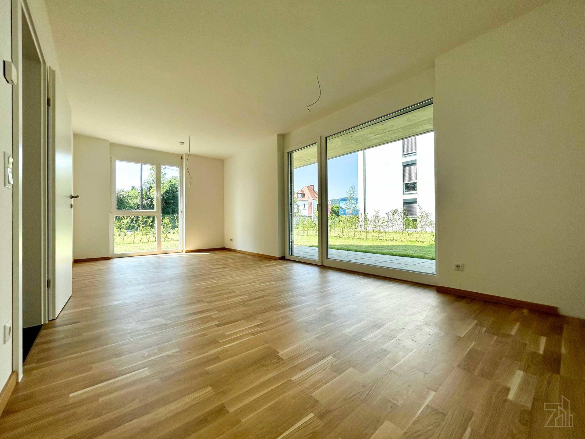 Heller, leerer Raum mit Parkettboden und großen Fenstern, die viel Tageslicht hereinlassen.