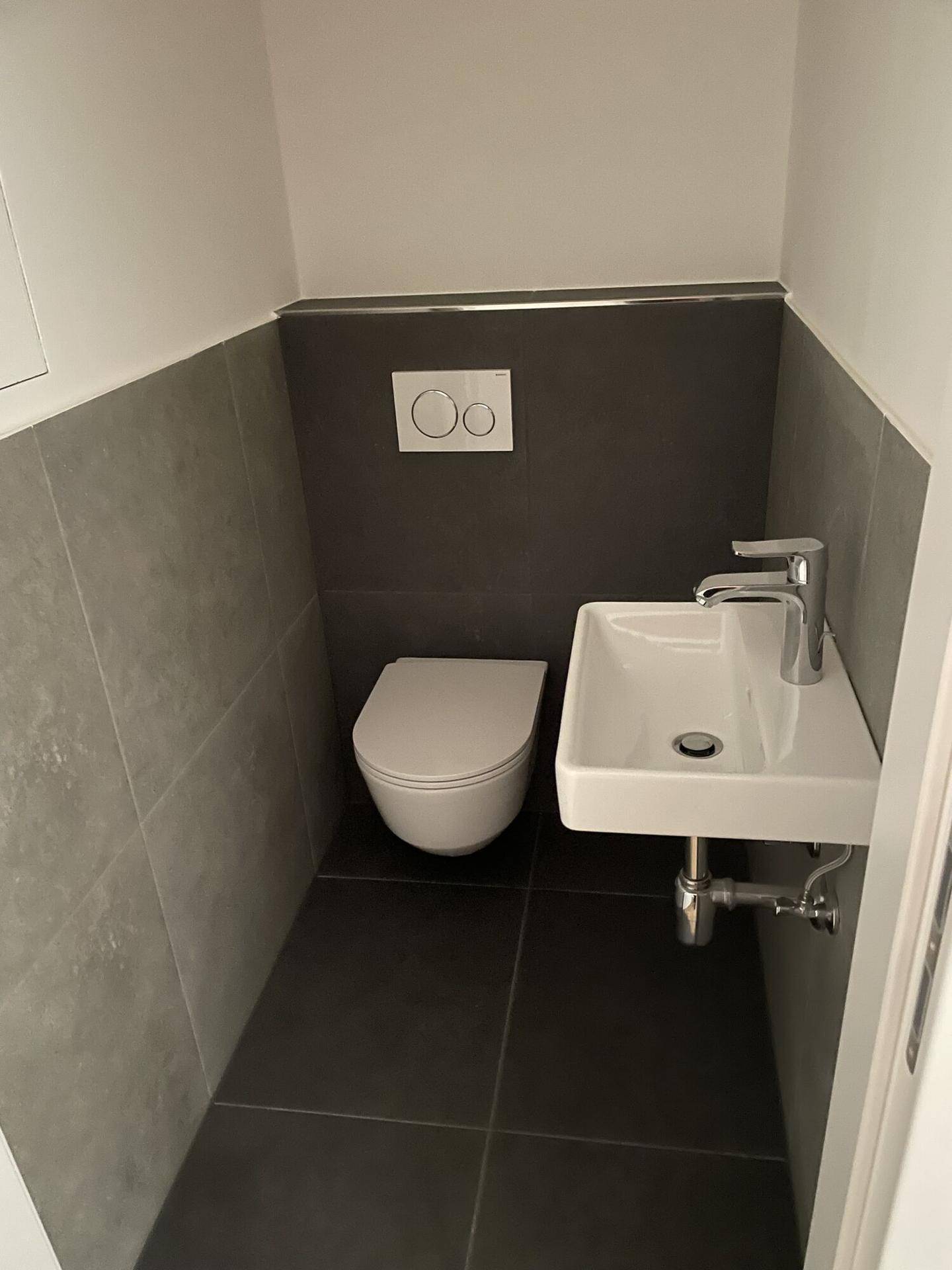 Modernes Gäste-WC mit wandhängender Toilette, kleinem Waschbecken und grauen Fliesen.