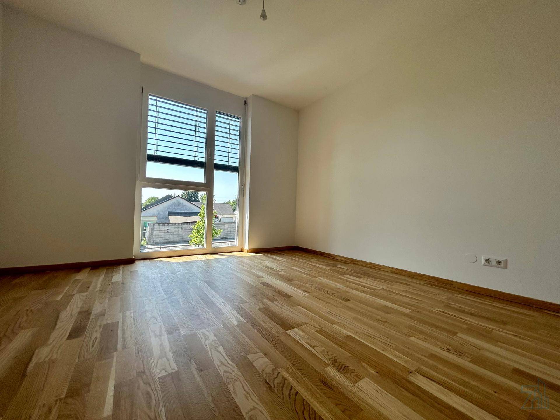 Lichtdurchflutetes Zimmer mit Holzboden und großem Fenster mit Jalousien.