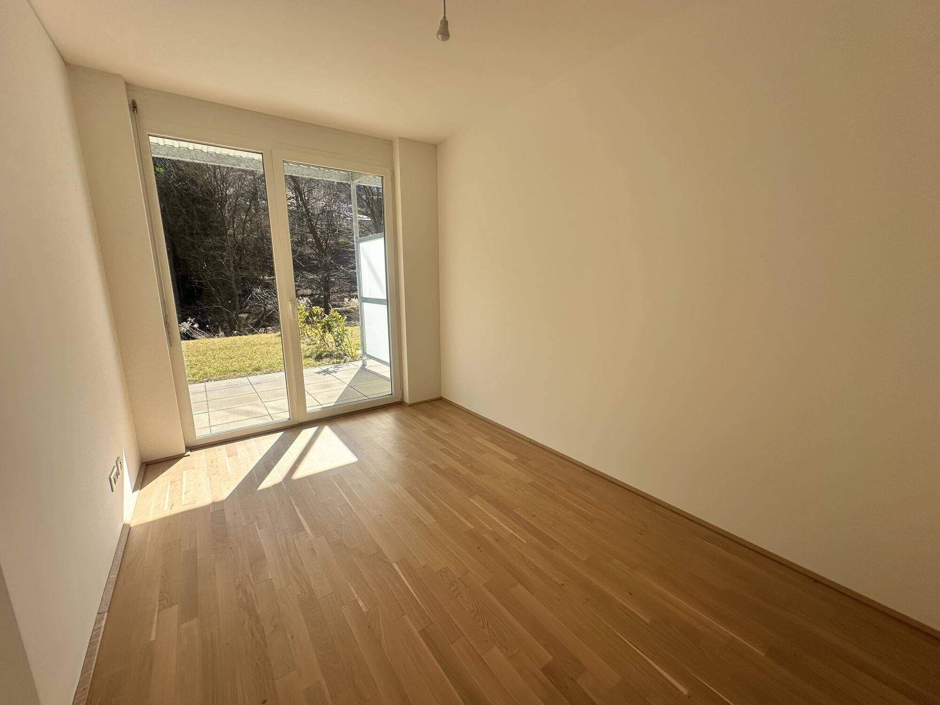 Leerer Raum mit helzernem Parkettboden und einer großen Glasschiebetür, die Zugang zu einem sonnigen Außenbereich bietet.