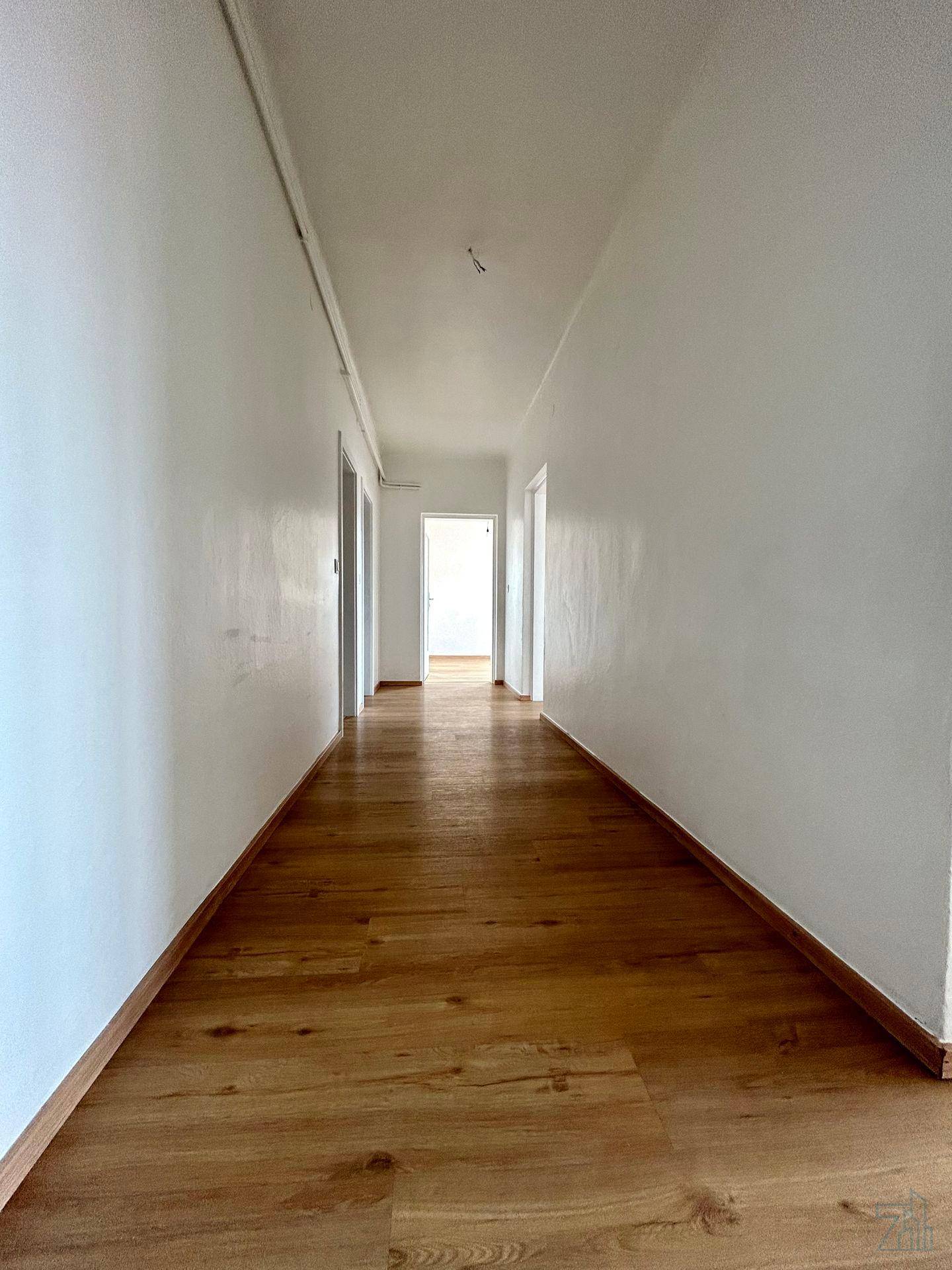 Langer, heller Flur mit Holzfußboden und weißen Wänden, der zu mehreren Zimmern führt.