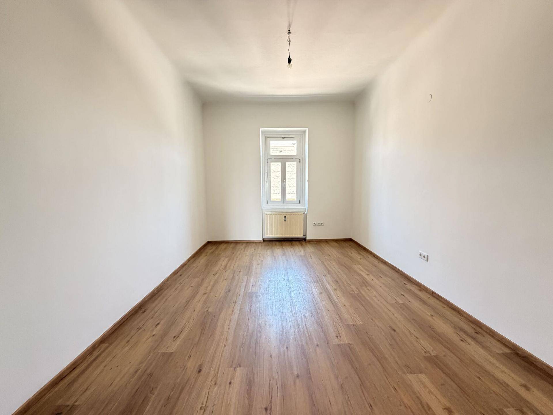 Leerer Raum mit Laminatboden, weißen Wänden und einem Fenster mit Heizkörper.