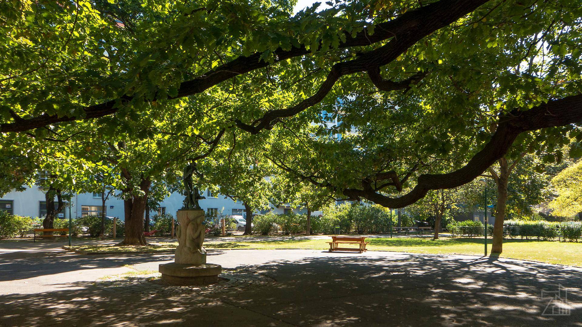 Sonniger Park mit alten Bäumen, Bänken und einer Statue, ideal zur Erholung.