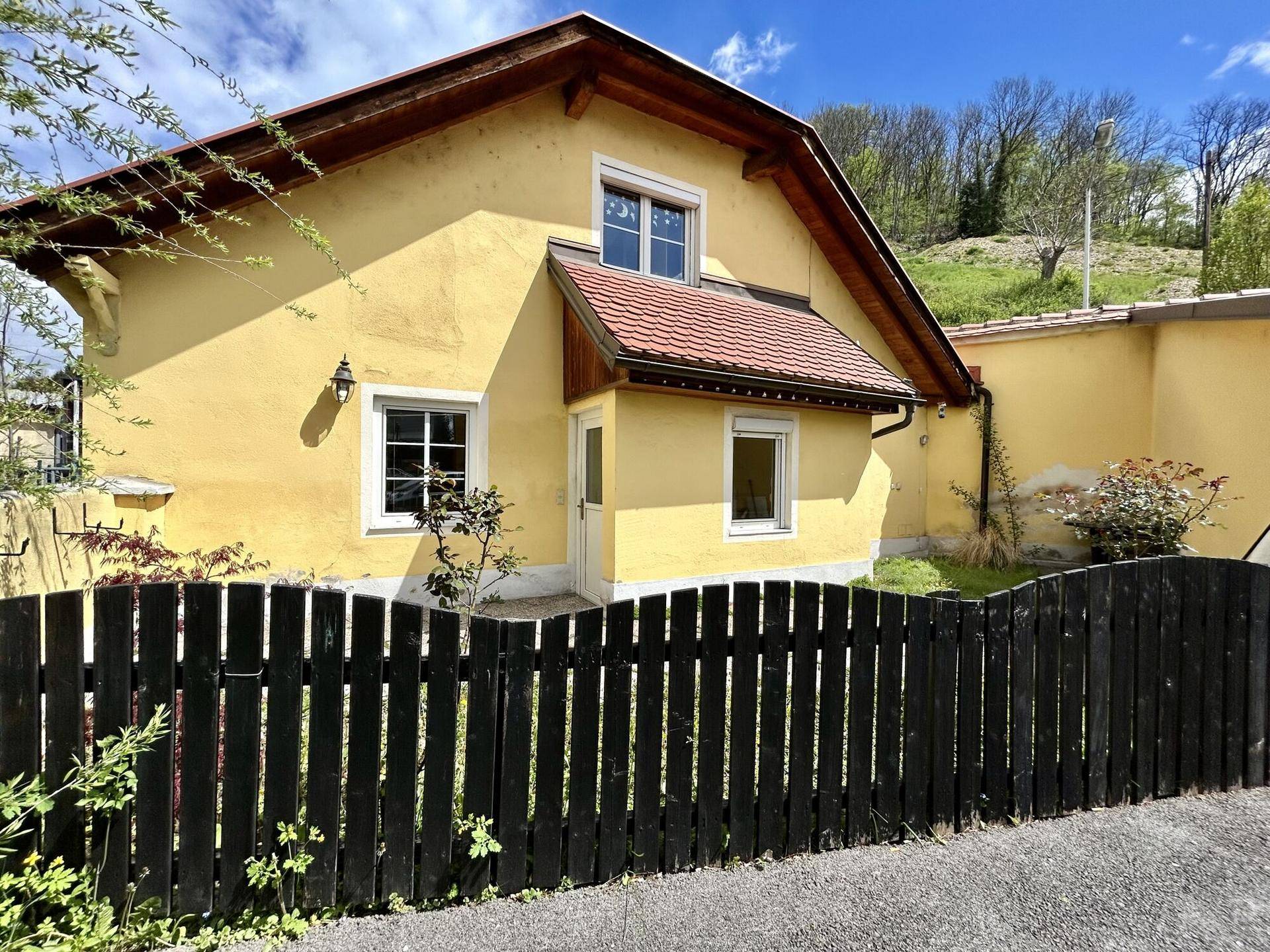 Seitenansicht des gelben Hauses mit schwarzem Holzzaun und grüner Umgebung.