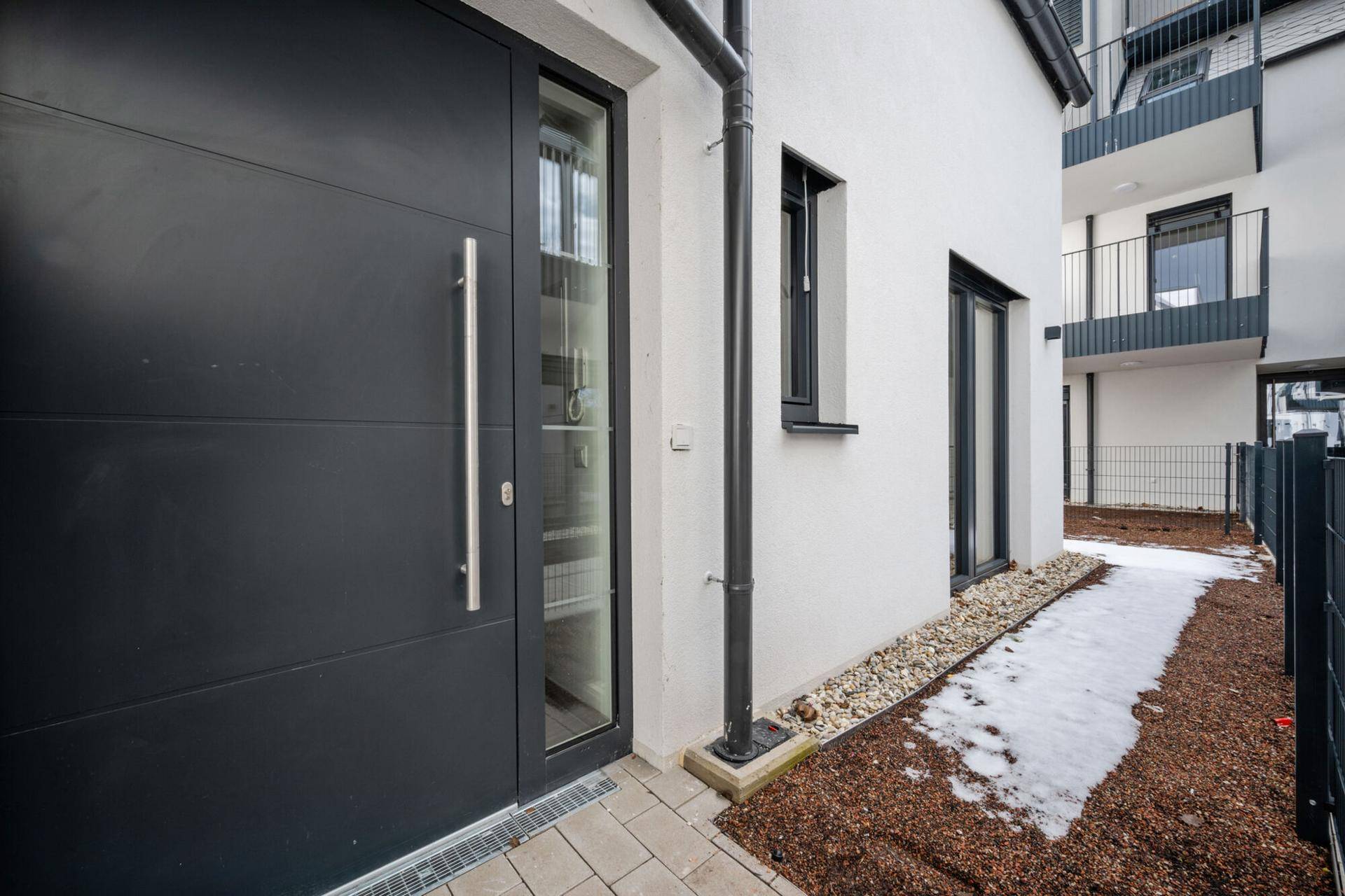 Moderner Hauseingang mit dunkler Tür und seitlichem Fenster, umgeben von Kies und Schnee.
