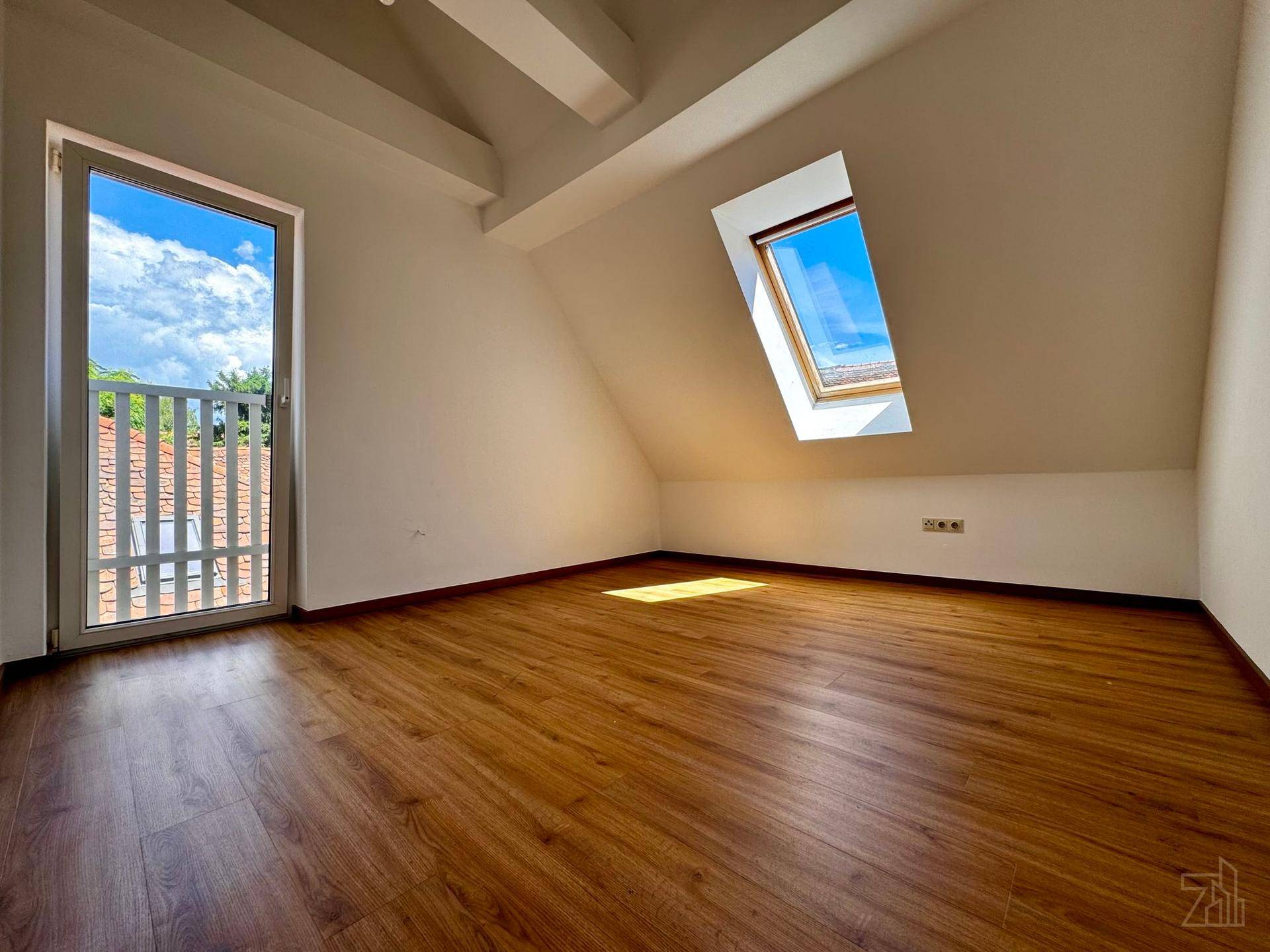 Helles Dachzimmer mit Holzboden, Dachschräge, Dachfenster und Zugang zu einem kleinen Balkon.
