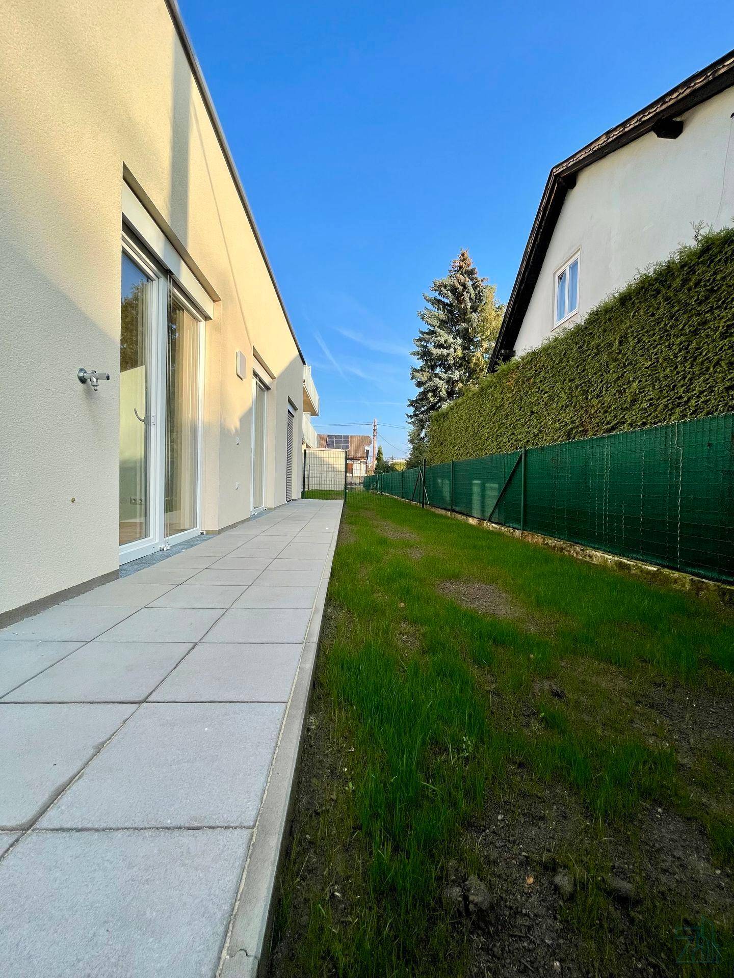 Seitenansicht des Gebäudes mit gepflasterter Terrasse und schmalem Rasenstreifen neben einer hohen Hecke.