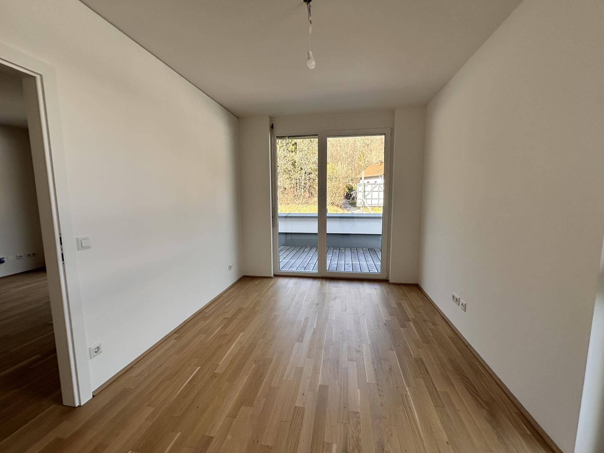 Geräumiger, heller Raum mit Holzboden und großen Fenstern, die ins Grüne blicken.