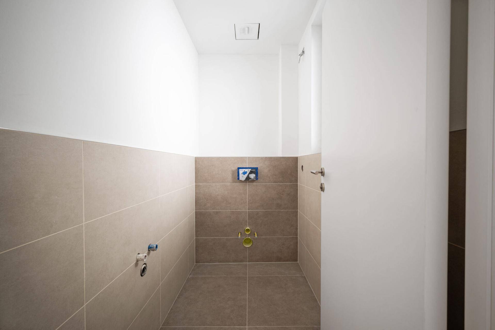 Kleiner WC-Raum mit beigefarbenen Fliesen und vorbereiteten Anschlüssen.