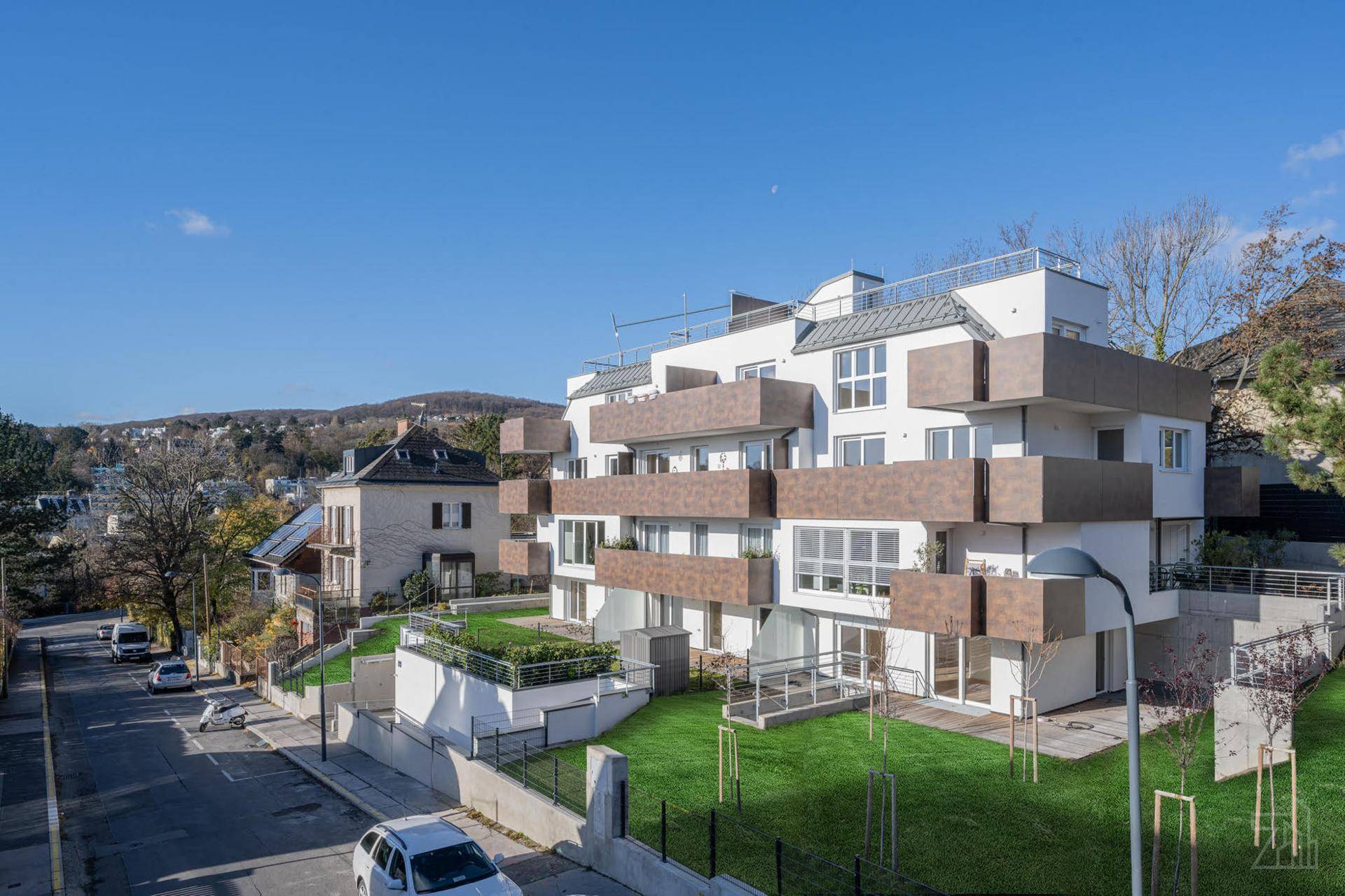 Modernes Mehrfamilienhaus mit mehreren Balkonen, begrünten Flächen und Blick auf die Straße.