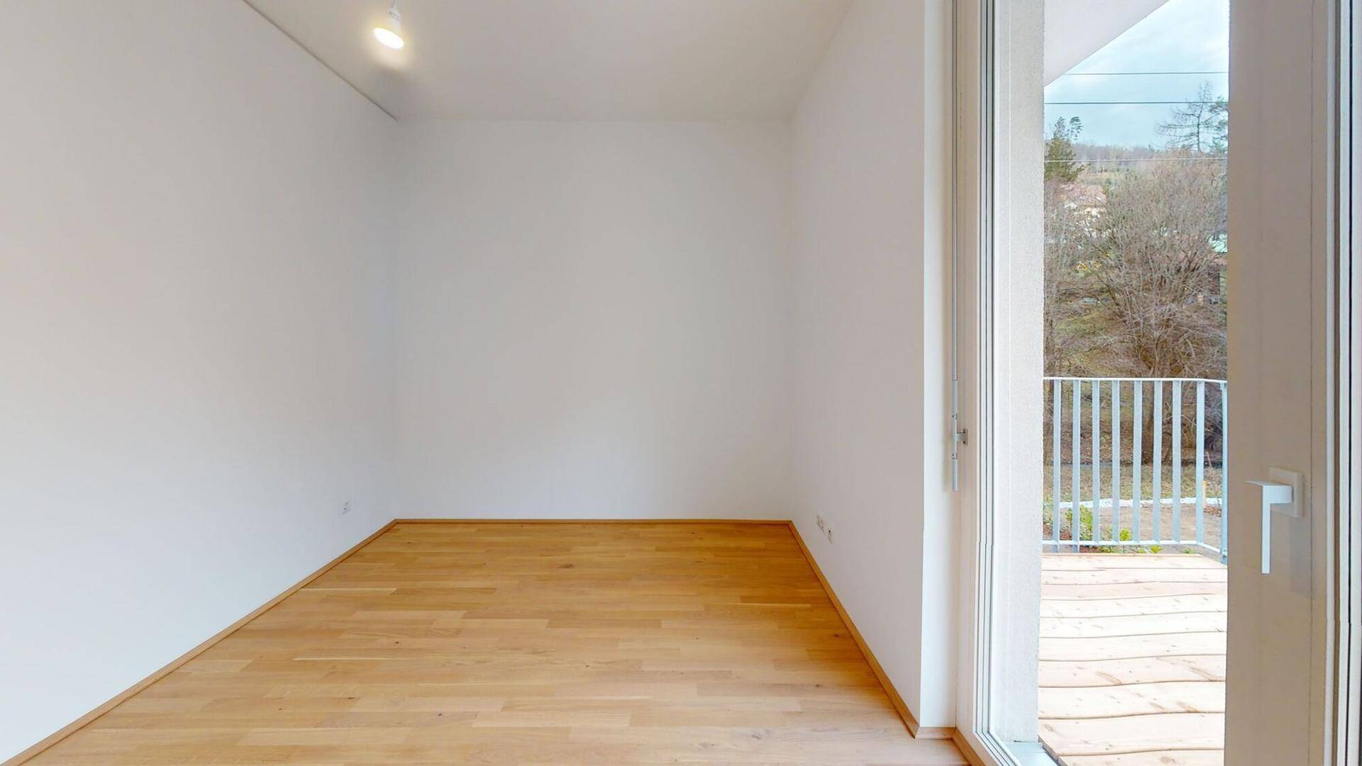 Leerer Raum mit hellem Holzboden und großer Balkontür, die viel Tageslicht hereinlässt.