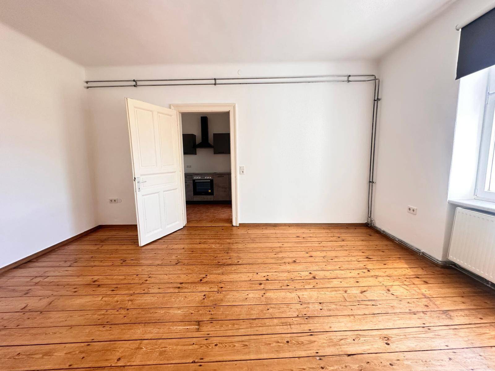 Geräumiger Wohnbereich mit Holzdielenboden und offener Tür zur angrenzenden Küche.