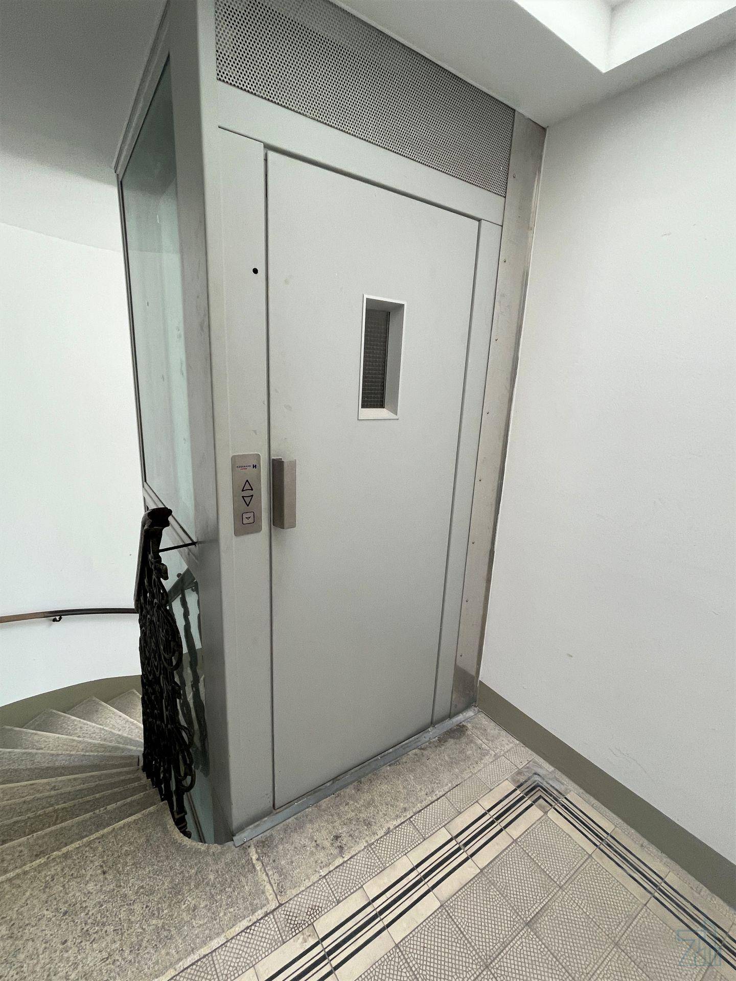 Kleiner Aufzug in einem Treppenhaus mit einer Wendeltreppe, zugänglich für alle Etagen.